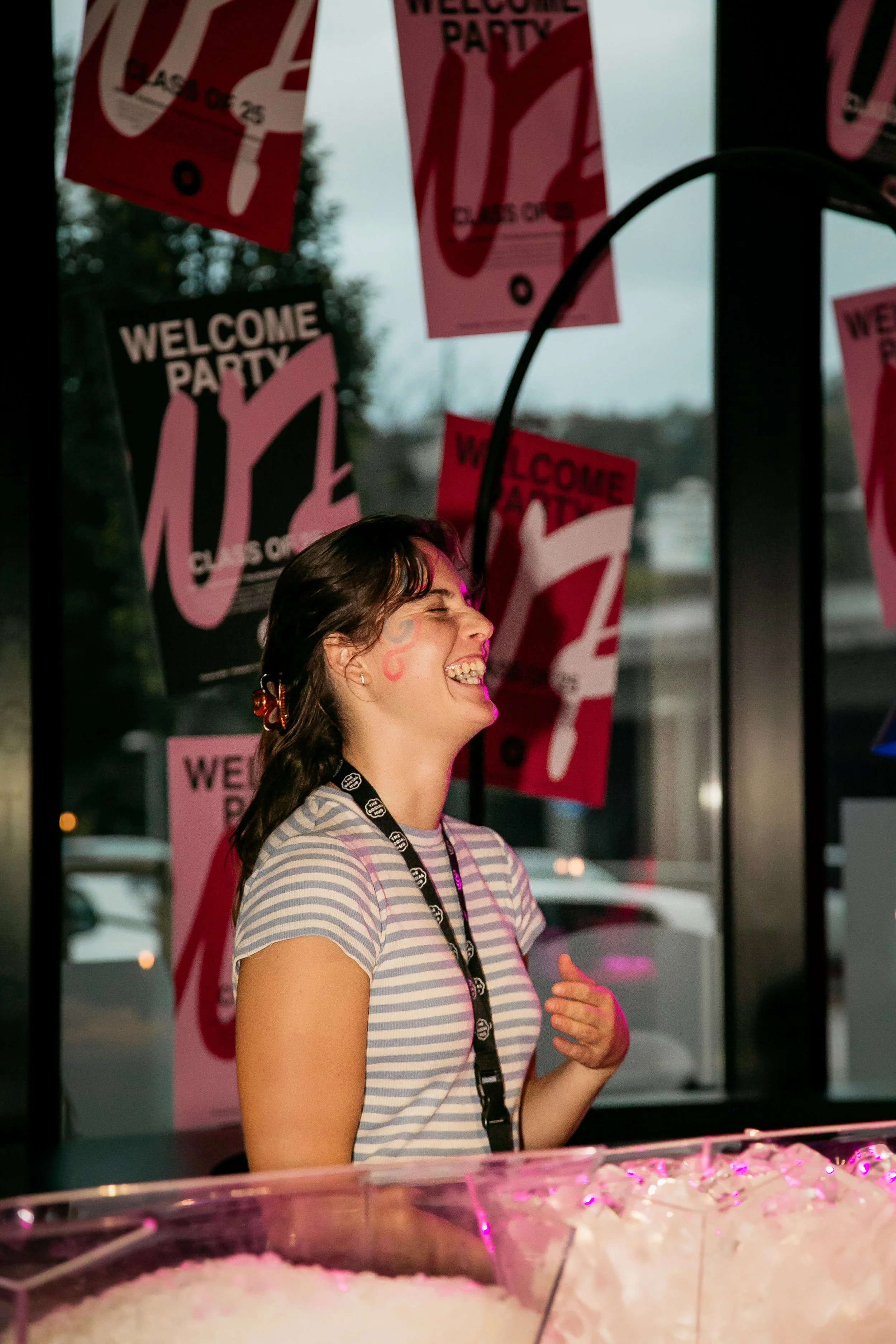 Joven sonriendo y hablando, con decoraciones de fiesta en el fondo que dicen 'Welcome Party' en colores rosa y negro.