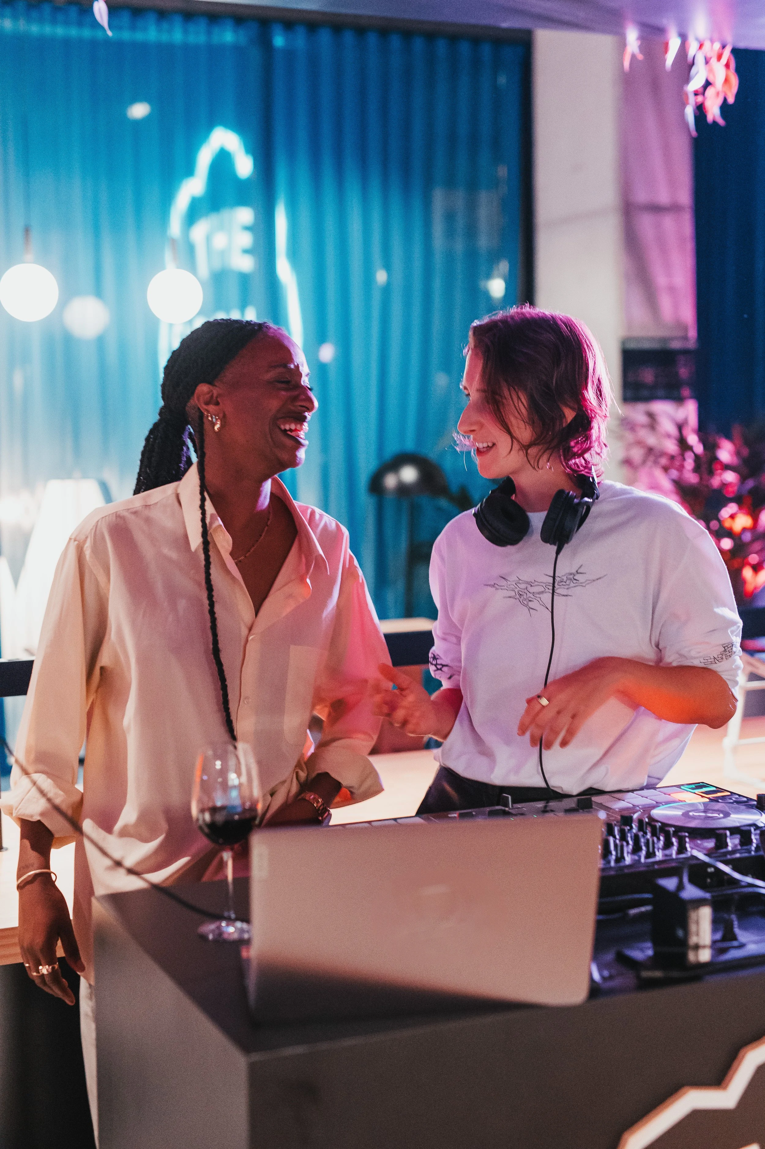 Dos mujeres en un ambiente de fiesta, una con cabello rizado y auriculares alrededor del cuello, la otra con cabello trenzado, compartiendo una sonrisa con fondo de cortinas y decoración luminosa.