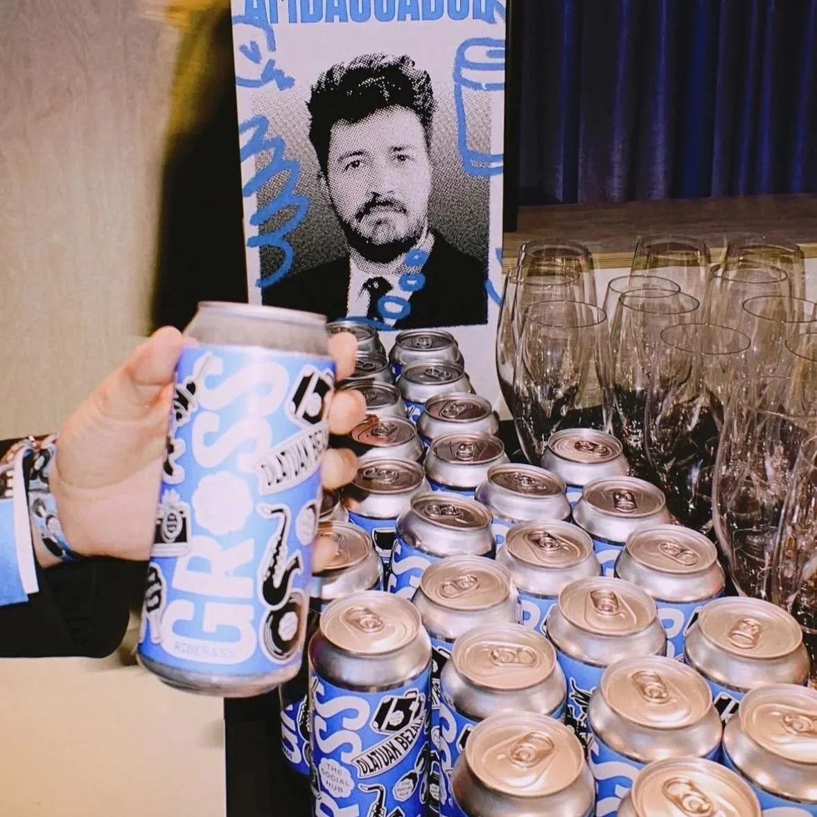 Mesa con muchas latas de bebidas, algunas copas vacías a la derecha y un cartel con la cara de un hombre. Persona sosteniendo una lata de bebida en primer plano.