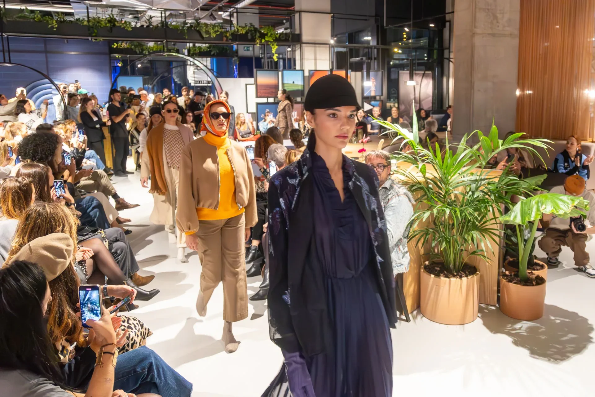 Modelos caminando por una pasarela en un evento de moda en un espacio interior decorado con plantas y cuadros en las paredes, mientras la audiencia las observa y toma fotos.