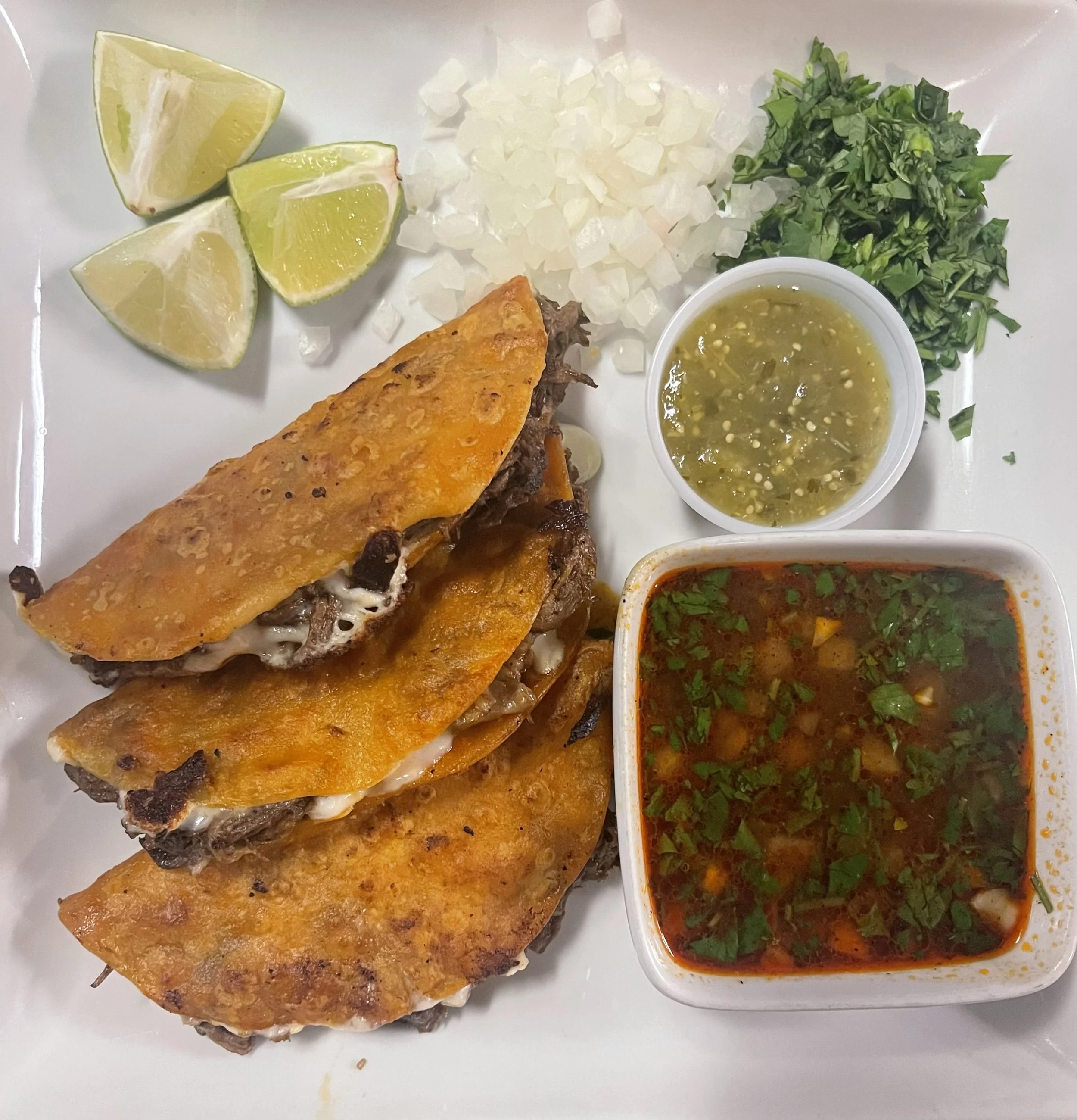 Plate with beef tacos topped with crispy cheese tortillas, lime wedges, chopped onions, chopped cilantro, salsa verde, and a bowl of spicy beef soup with herbs.