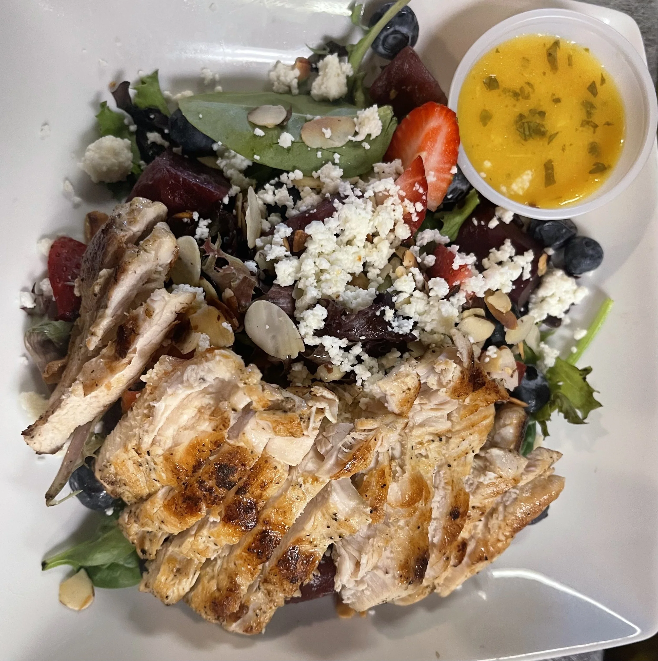 Salad with sliced grilled chicken, mixed greens, strawberries, blueberries, beets, feta cheese, sliced almonds, pumpkin seeds, and a side of vinaigrette dressing.