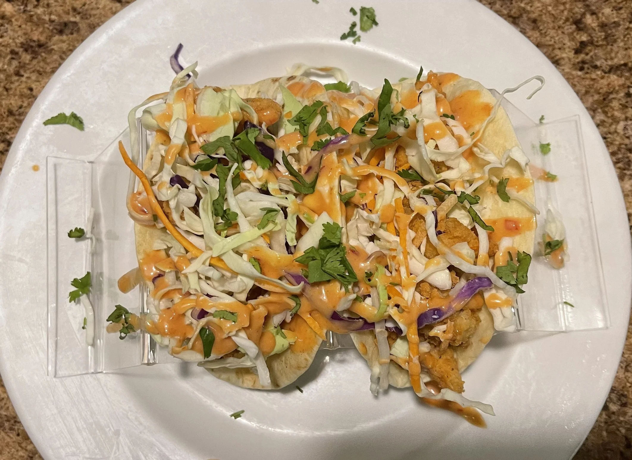 Two fish tacos topped with shredded lettuce, cilantro, and drizzled with orange sauce on a white plate.