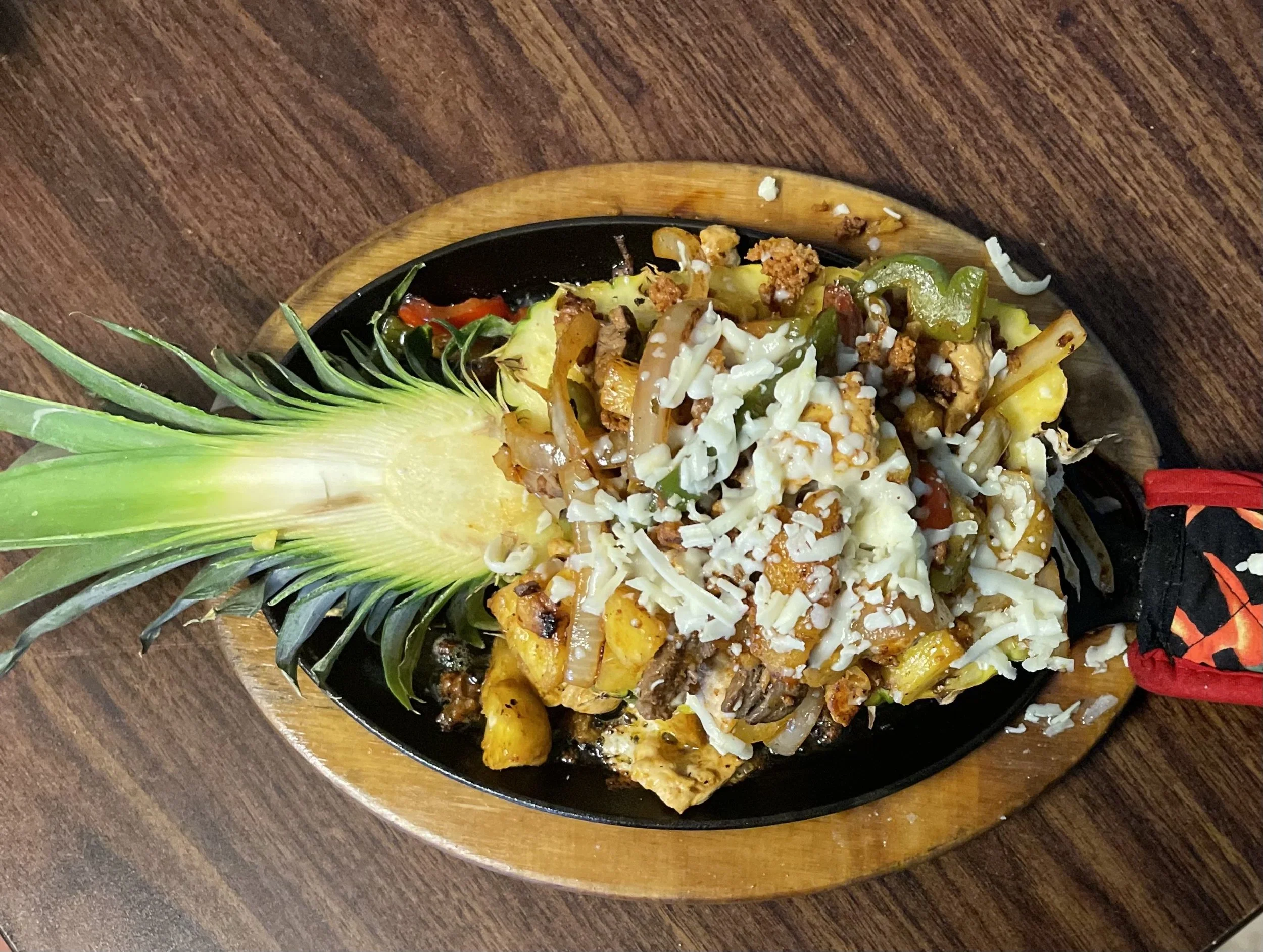 Pineapple shaped serving of Mexican fajitas with grilled vegetables, shredded cheese, and pineapple leaves, served on a black sizzling plate with a wooden base on a dark wooden table.