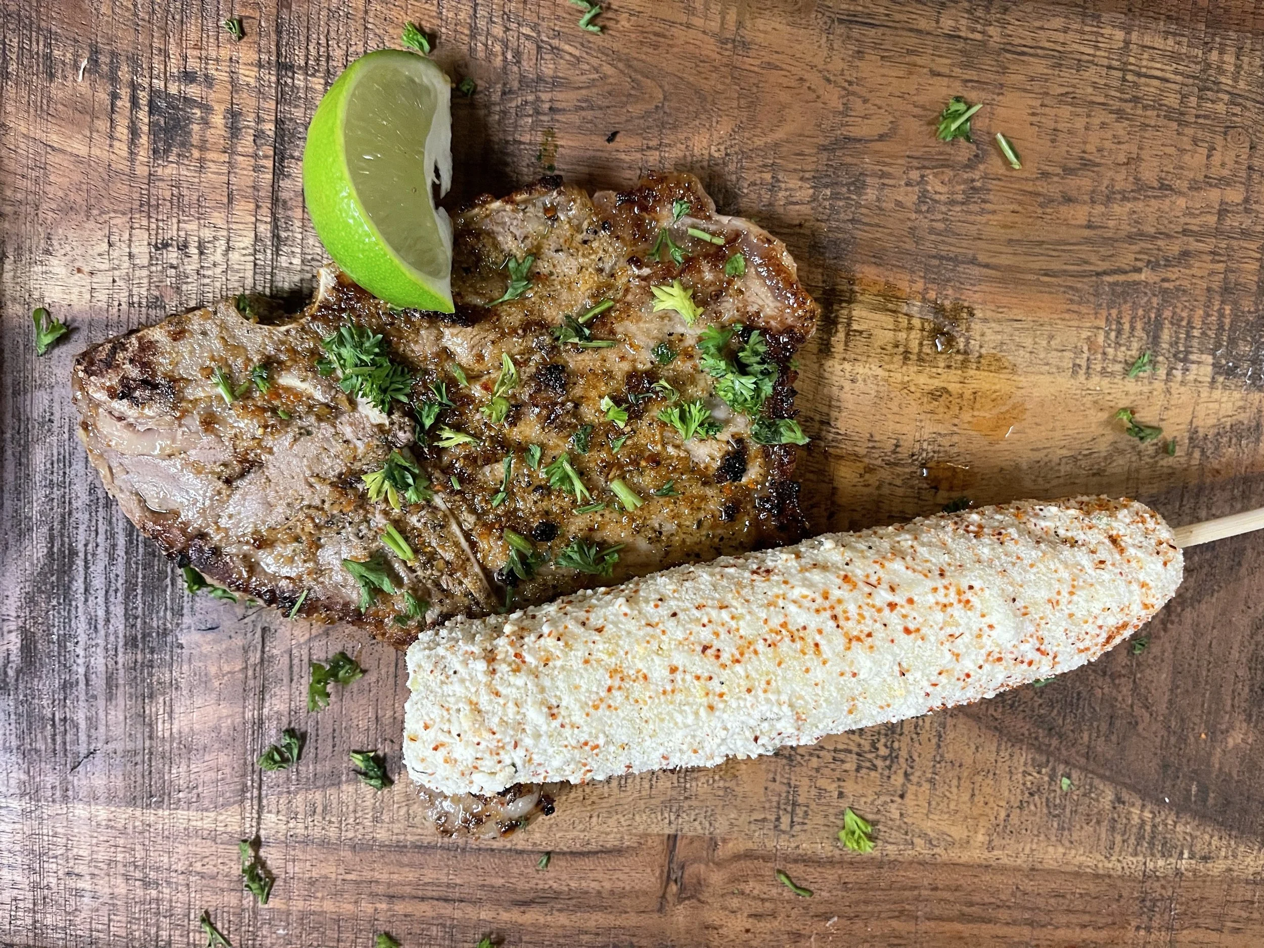 Grilled steak garnished with chopped herbs, accompanied by a wedge of lime and Mexican corn on a wooden cutting board.