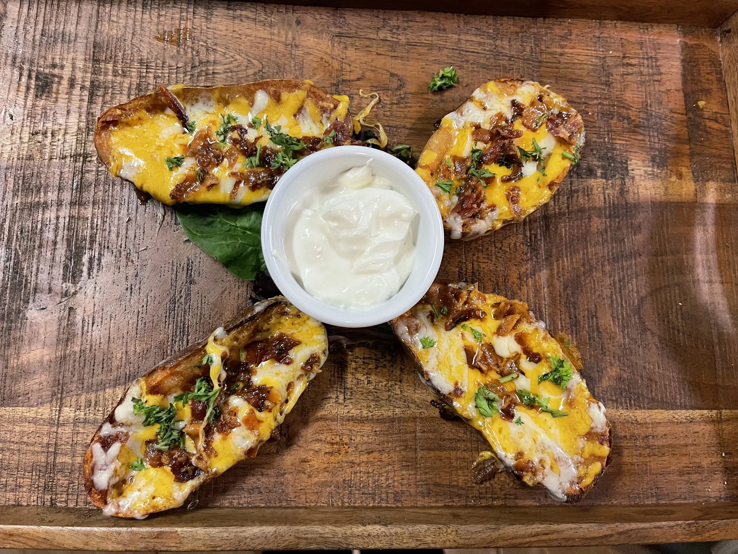 Four loaded potato skins with cheese, chopped herbs, arranged in a circle around a bowl of white dipping sauce on a wooden serving board.