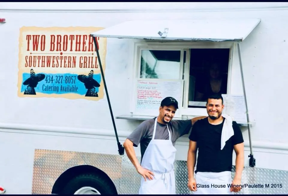 Two men smiling and standing outside a food truck called 'Two Brothers Southwestern Grill,' with one man wearing an apron and the other with a towel over his shoulder, in front of a window menu.