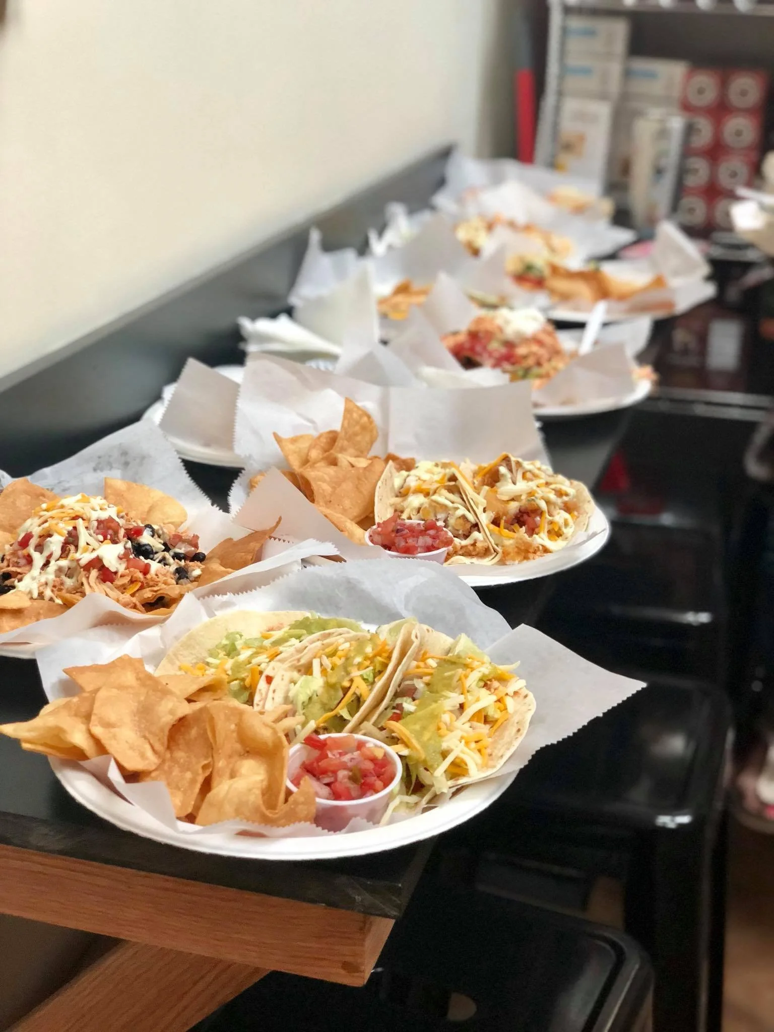 Multiple plates of tacos with tortilla shells, shredded cheese, lettuce, and salsa, along with side chips and salsa, on a counter in a restaurant setting.