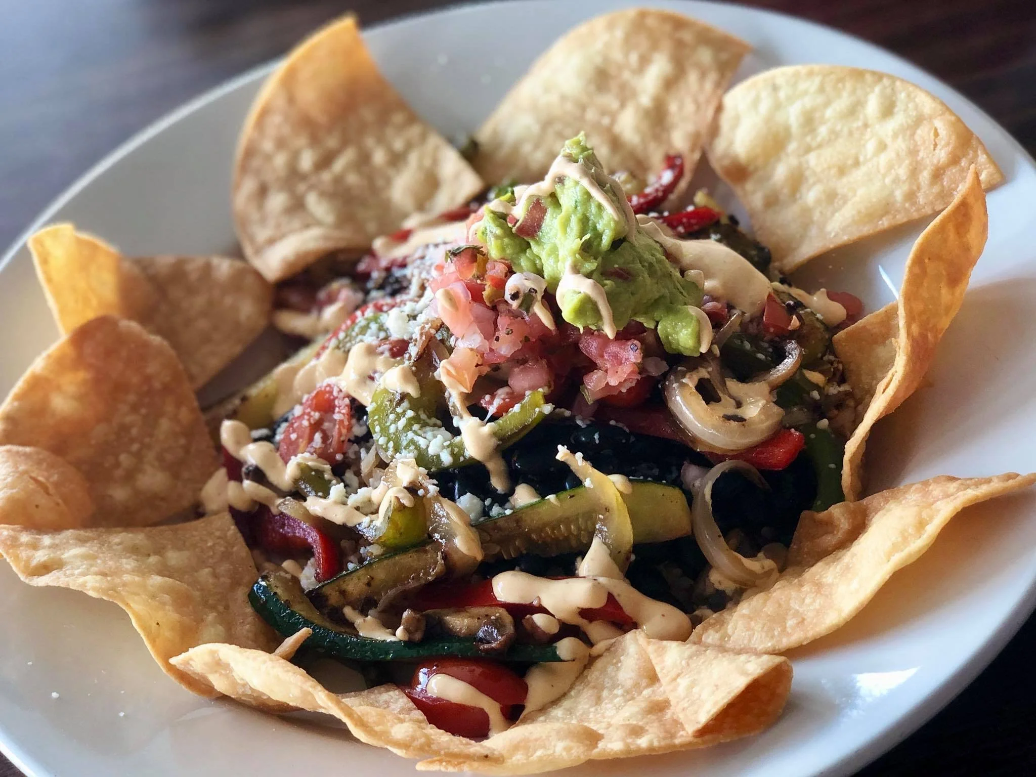 A plate of nachos topped with guacamole, sour cream, diced tomatoes, onions, peppers, olives, and shredded cheese, with tortilla chips around the edges.