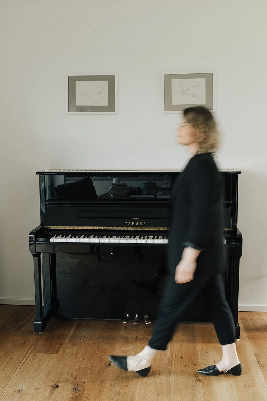 Julia in schwarzer Kleidung und schwarzen Schuhen geht vor einem schwarzen Yamaha Klavier vorbei, das an einer weißen Wand steht. Über dem Klavier hängen zwei einfache, silberne Rahmen mit Kunstdrucken.