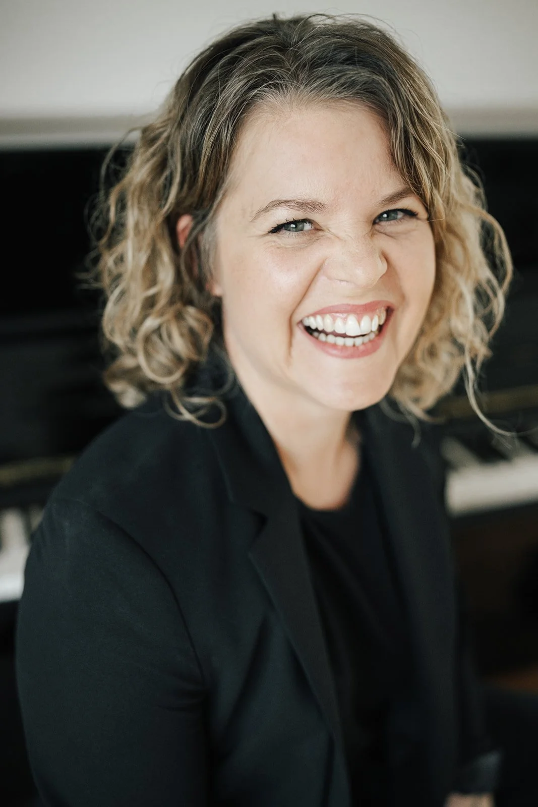 Eine lachende Frau mit curly Haaren trägt schwarze Kleidung und sitzt vor einem Klavier.