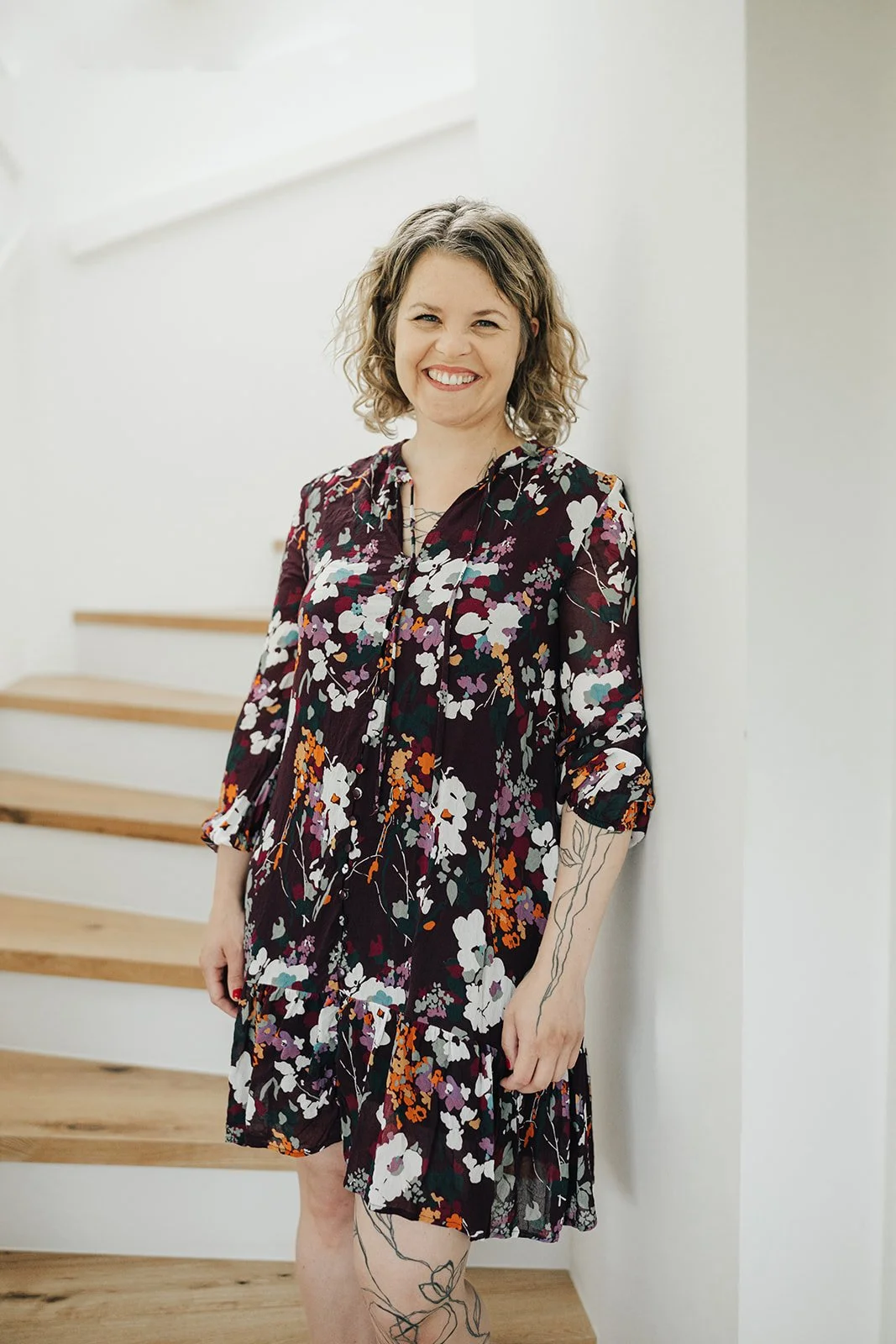 Eine lächelnde Frau mit lockigem Haar in einem floral gemusterten Kleid, die an einer weißen Wand steht.