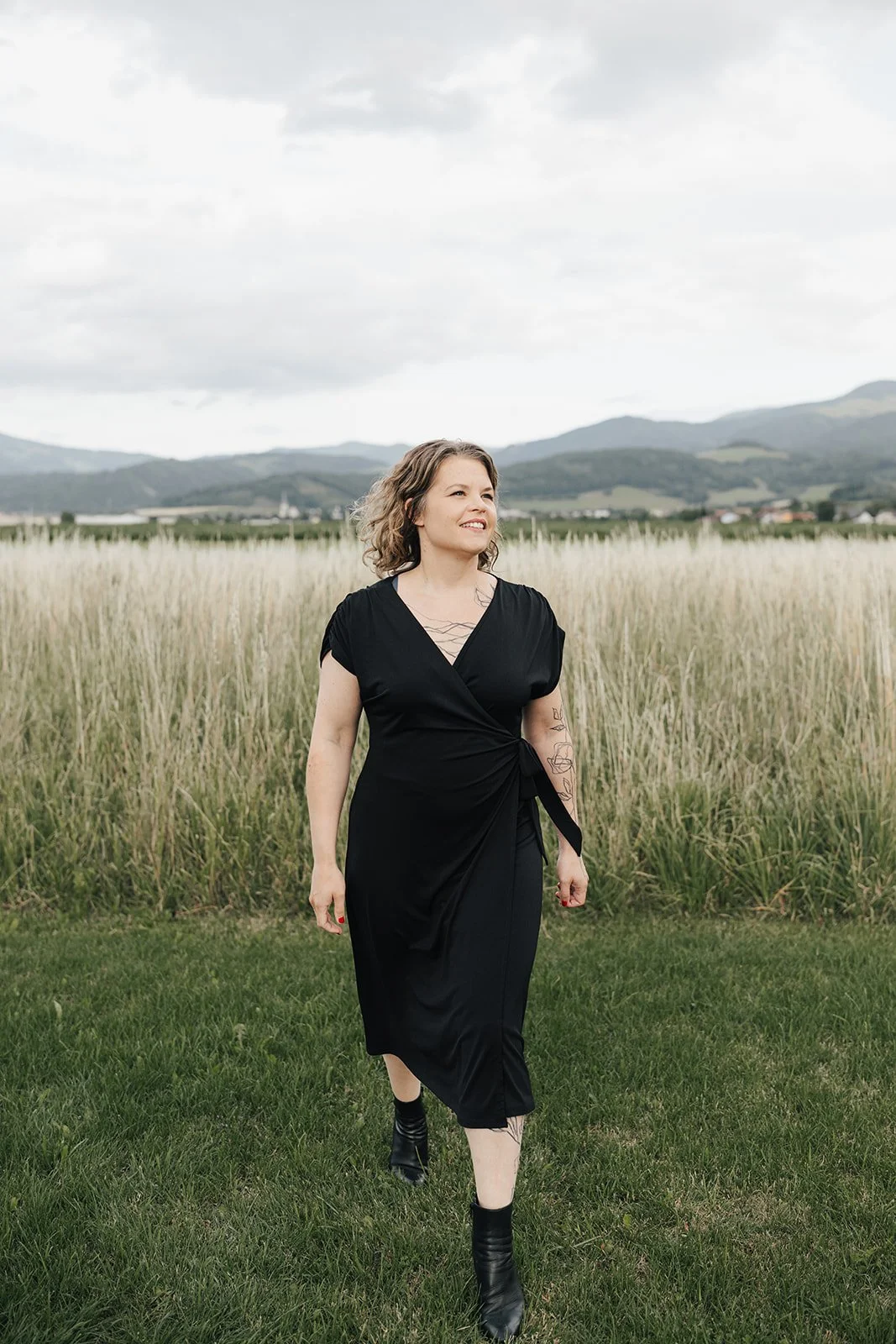 Junge Frau in schwarzem Kleid läuft durch grüne Wiese mit hohen Gräsern, im Hintergrund Hügel und Wolken am Himmel.