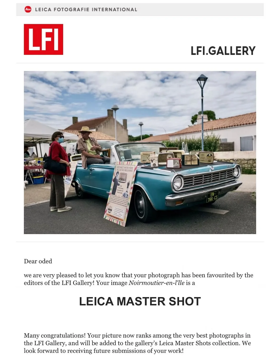 Une voiture rétro bleu exposée lors d'une exposition, avec deux personnes discutant à côté, sous un ciel partiellement nuageux. A l'île de Noirmoutier, France.