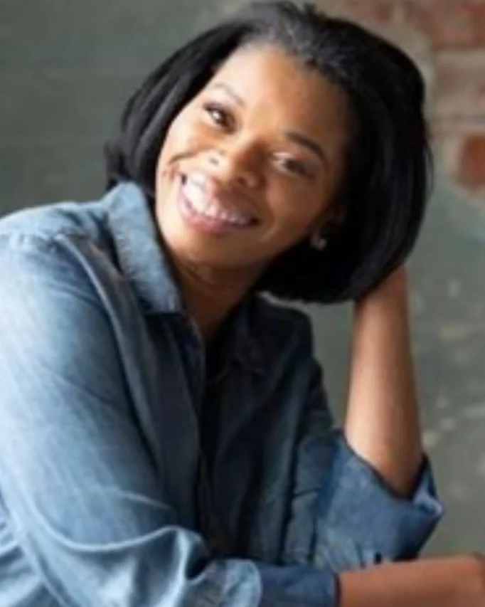 Smiling woman with black hair, wearing a denim shirt, sitting with her hand resting on her head in a casual setting.