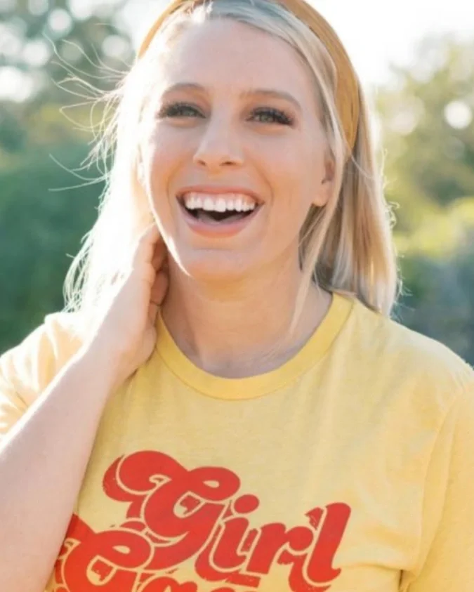 A woman smiling outdoors, wearing a yellow T-shirt with red text, and a yellow headband with sunlight and greenery in the background.