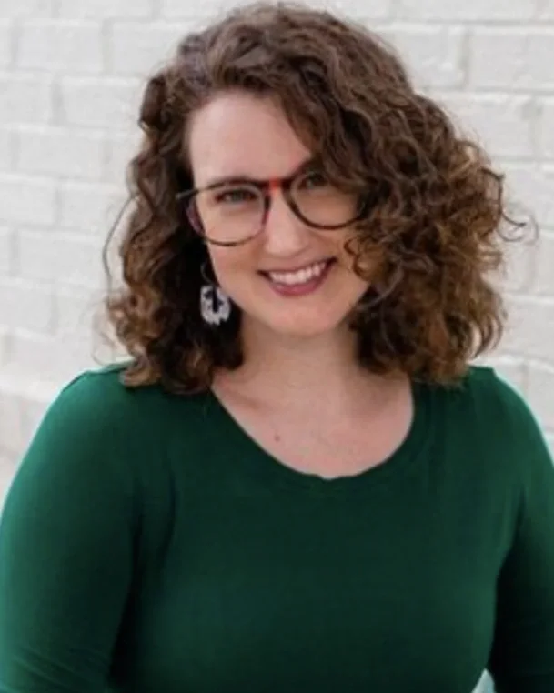 A woman with curly brown hair and glasses, wearing a green top, smiling in front of a brick wall.