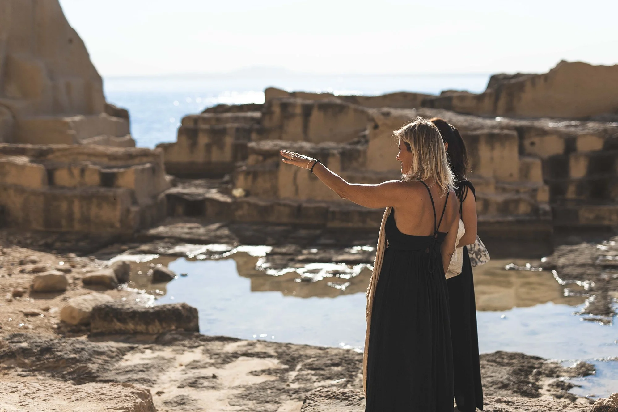 Two women standing on rocky shores by the ocean, one with blonde hair in a black dress, with the ocean and cliffs in the background.