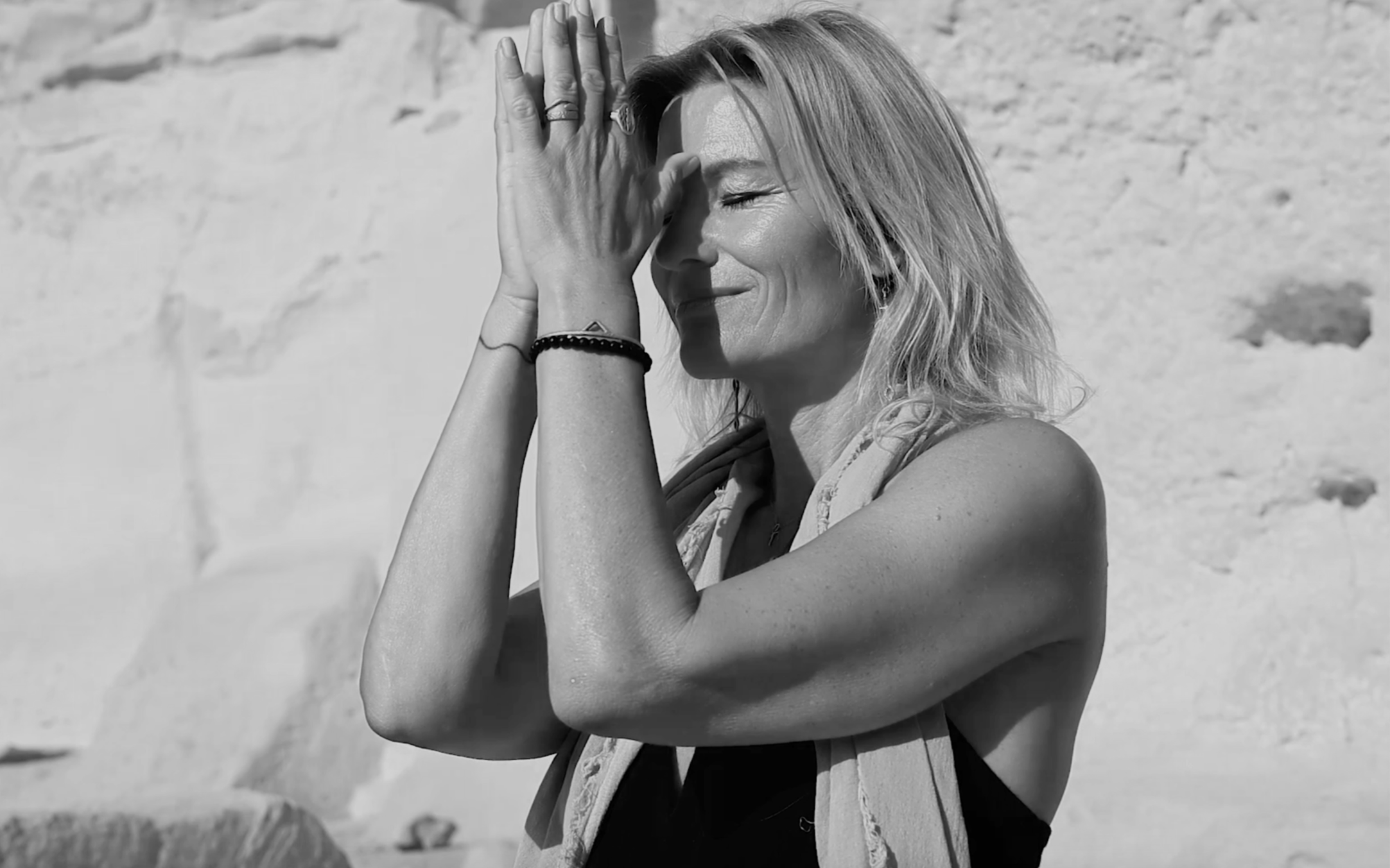 A woman with blonde hair, closing her eyes and smiling, standing outdoors against a rocky background, with her hands pressed together near her forehead.