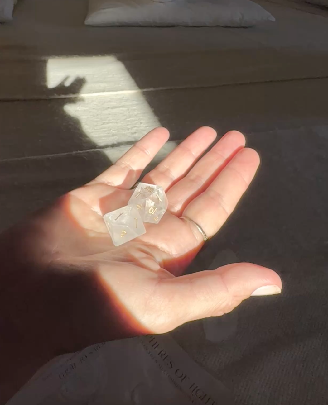 A person's hand holding two transparent dice with gold numbers, with sunlight casting a shadow and a blurred book in the background.