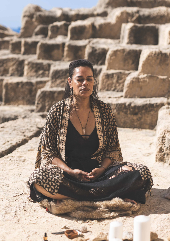 A woman sits cross-legged outdoors in front of large stone steps, meditating with her eyes closed, wearing a patterned shawl and black clothing, with candles and objects in front of her.