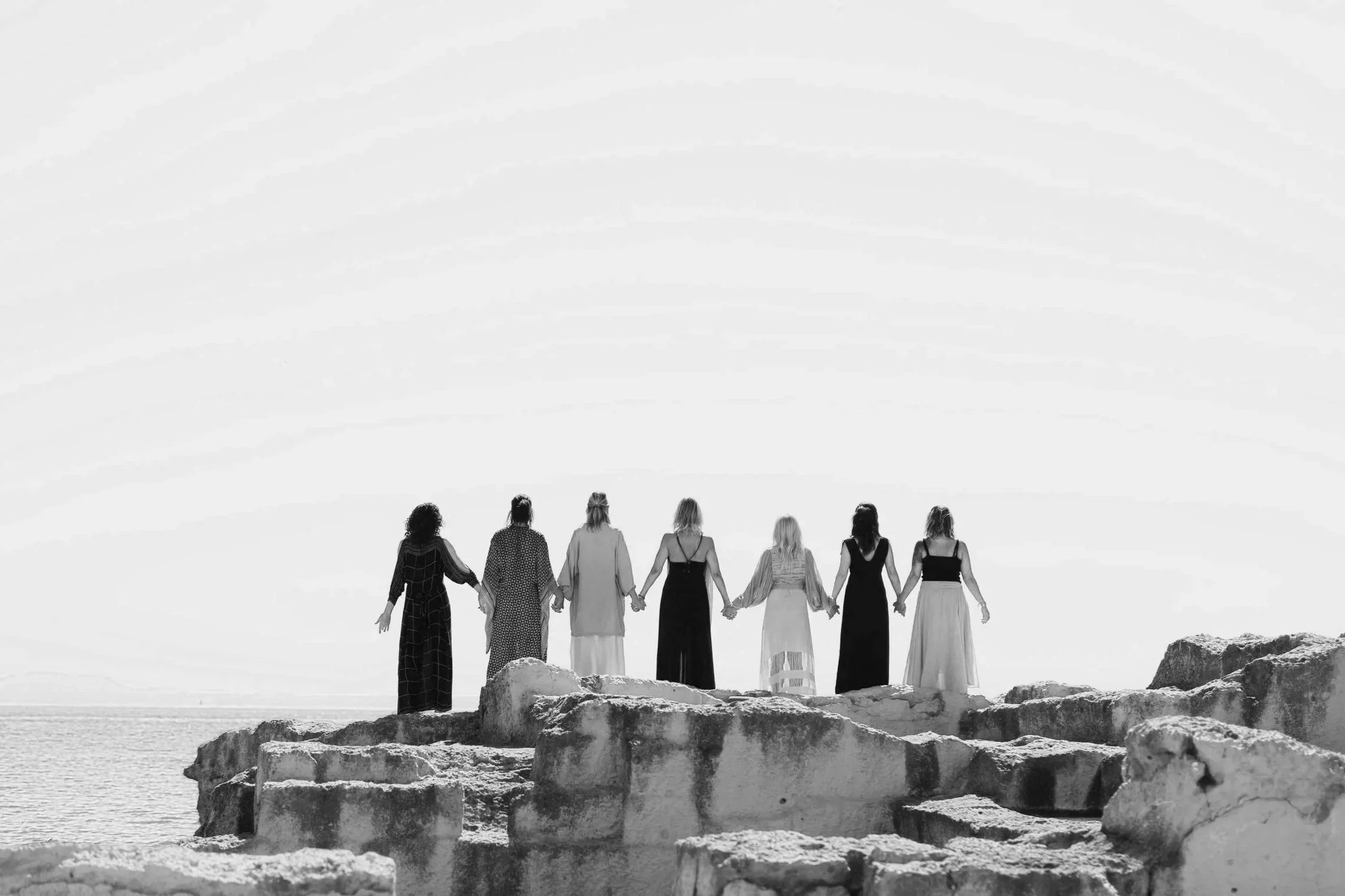 A group of women standing hand in hand on rocks by the sea, facing the ocean, in black and white.