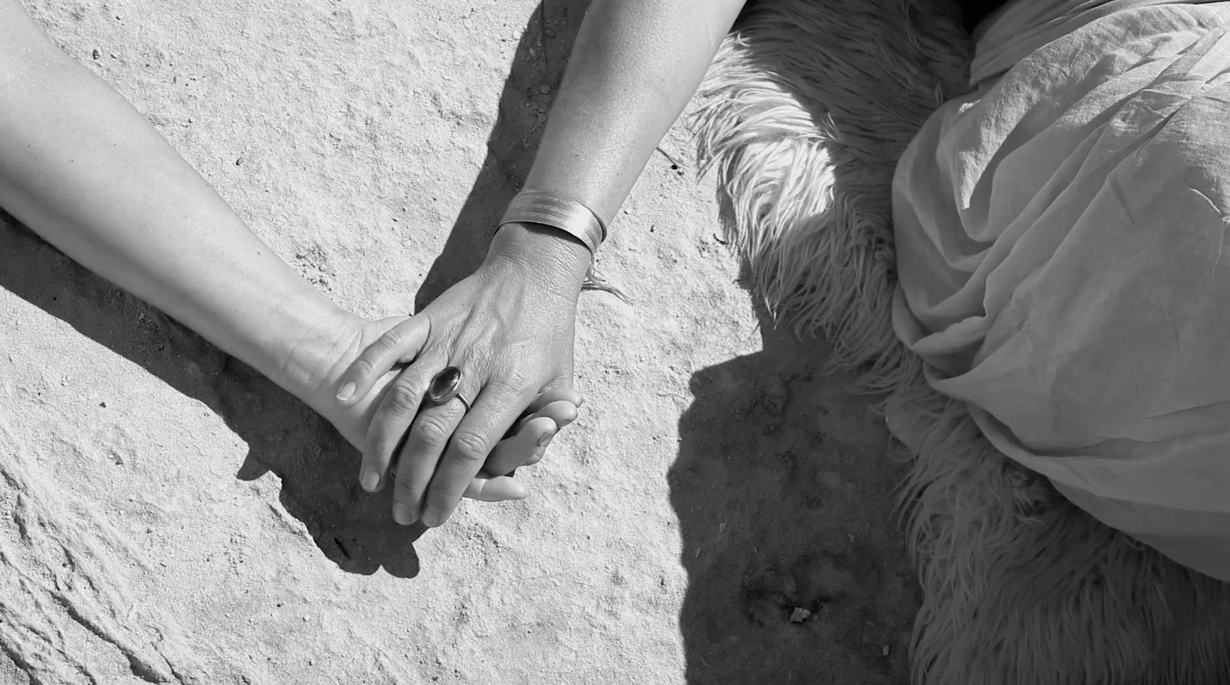 Two people holding hands, one person on the ground and the other kneeling, with sand or dirt beneath them.