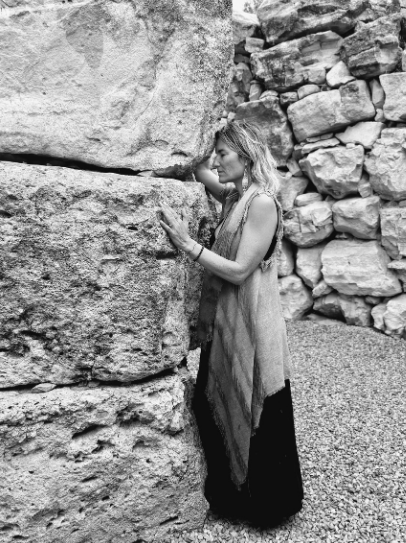 A woman with long hair, wearing a sleeveless dress and shawl, stands close to a large stone wall, lovingly touching and embracing it, with a background of other stone formations.