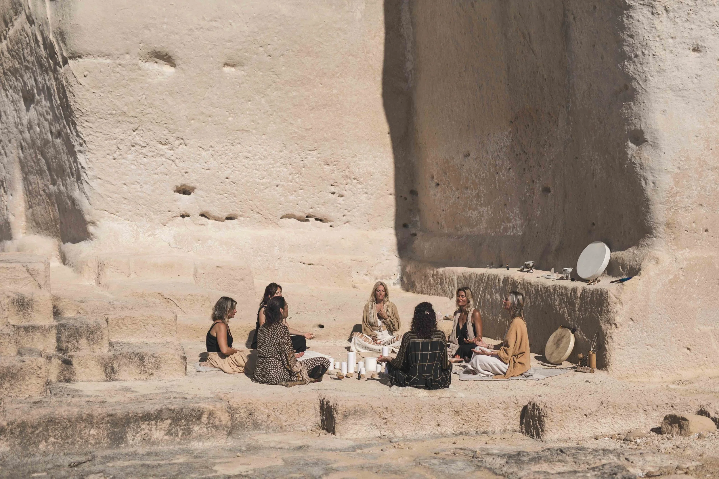 A group of women seated in a circle on steps in front of a large beige rock face, participating in a meditation or spiritual gathering.