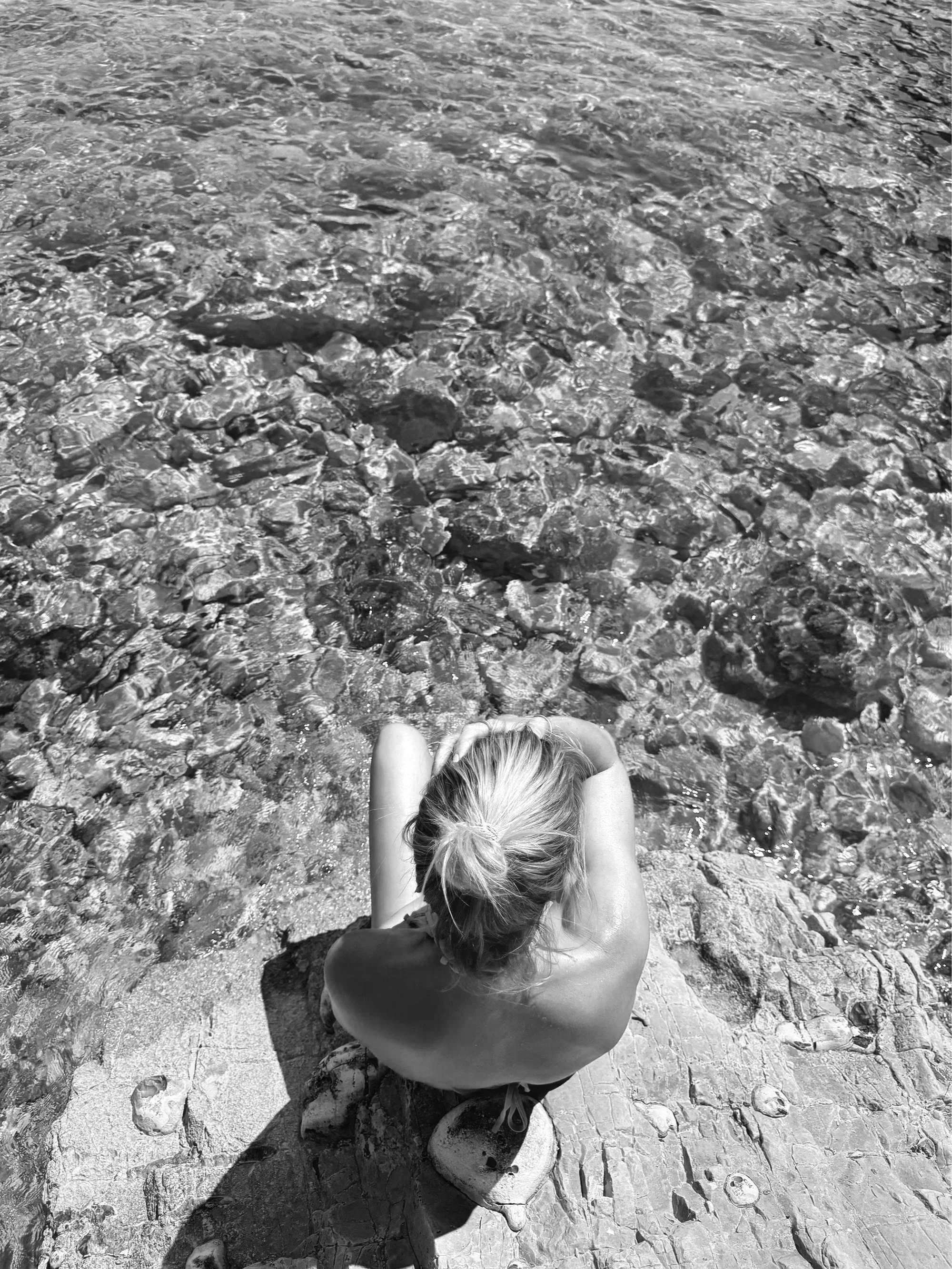 A woman with light-colored hair in a bun, sitting on a rocky shoreline with her arms resting on her knees, facing a body of water with pebbles and rocks visible beneath the surface.