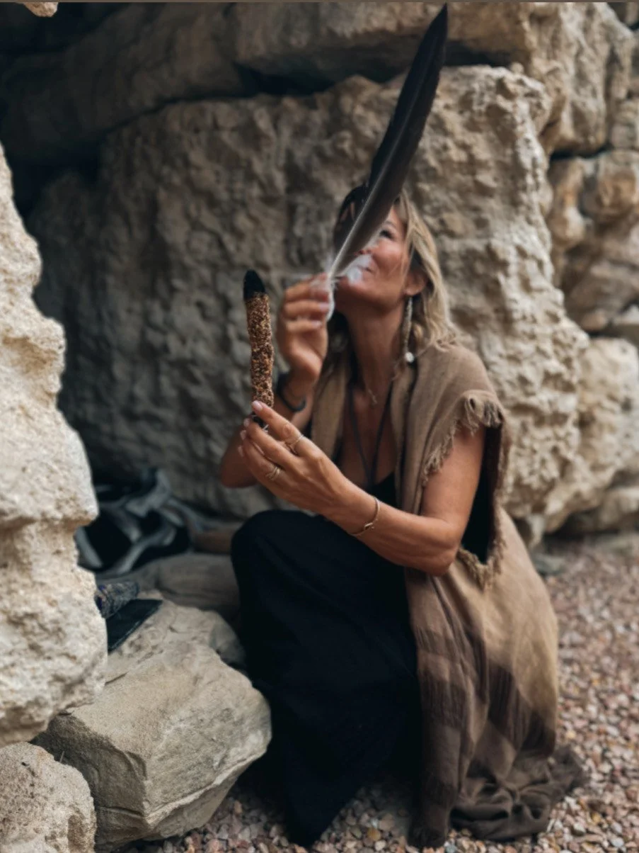 A woman with long blonde hair and bohemian style sitting among large rocks, holding a feather and a stick, with her face partially obscured by the feather.