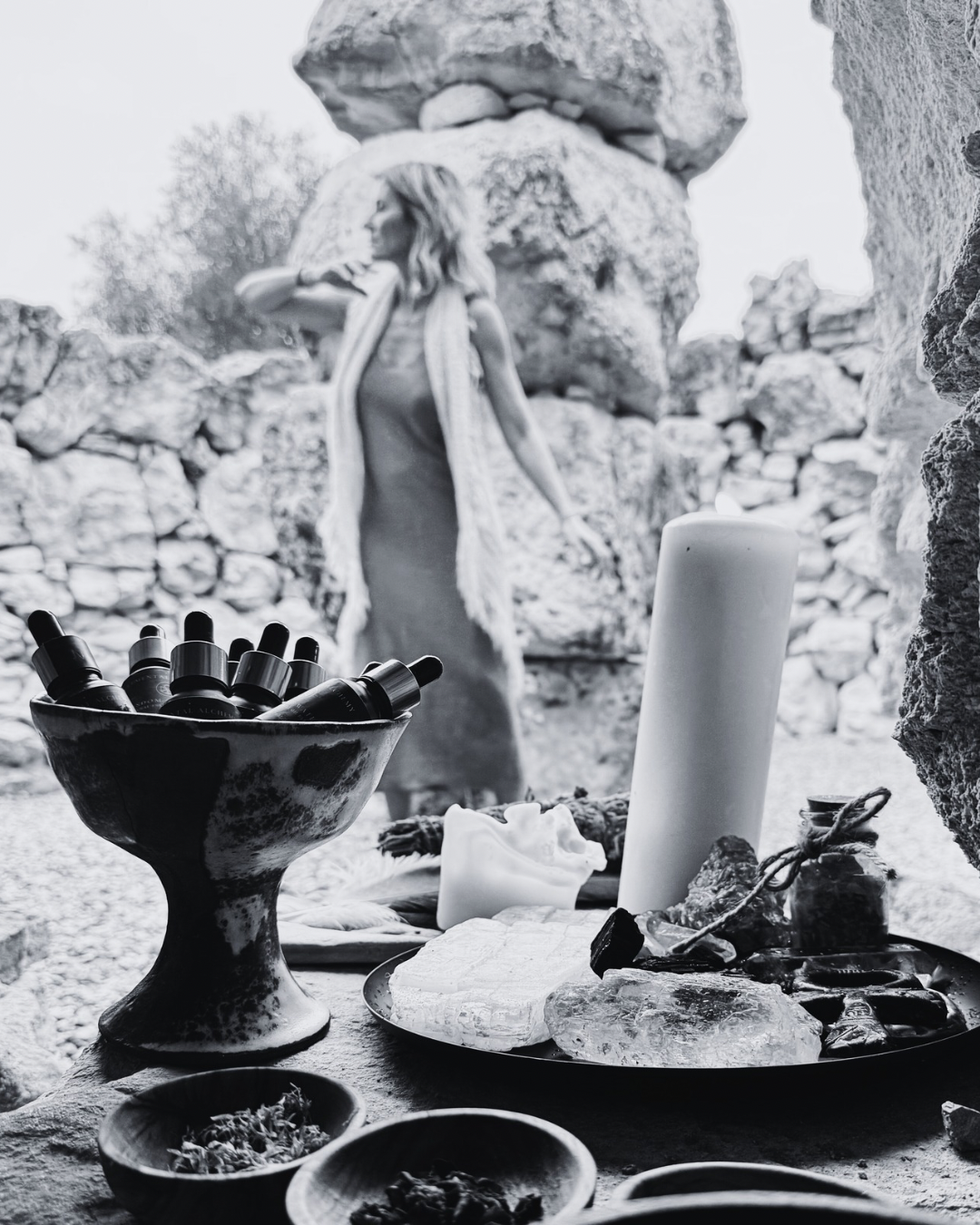 A woman in a sleeveless dress and scarf stands outdoors near a stone wall, with a table in the foreground containing candles, essential oils, rocks, and small bowls, creating a serene, spiritual atmosphere.