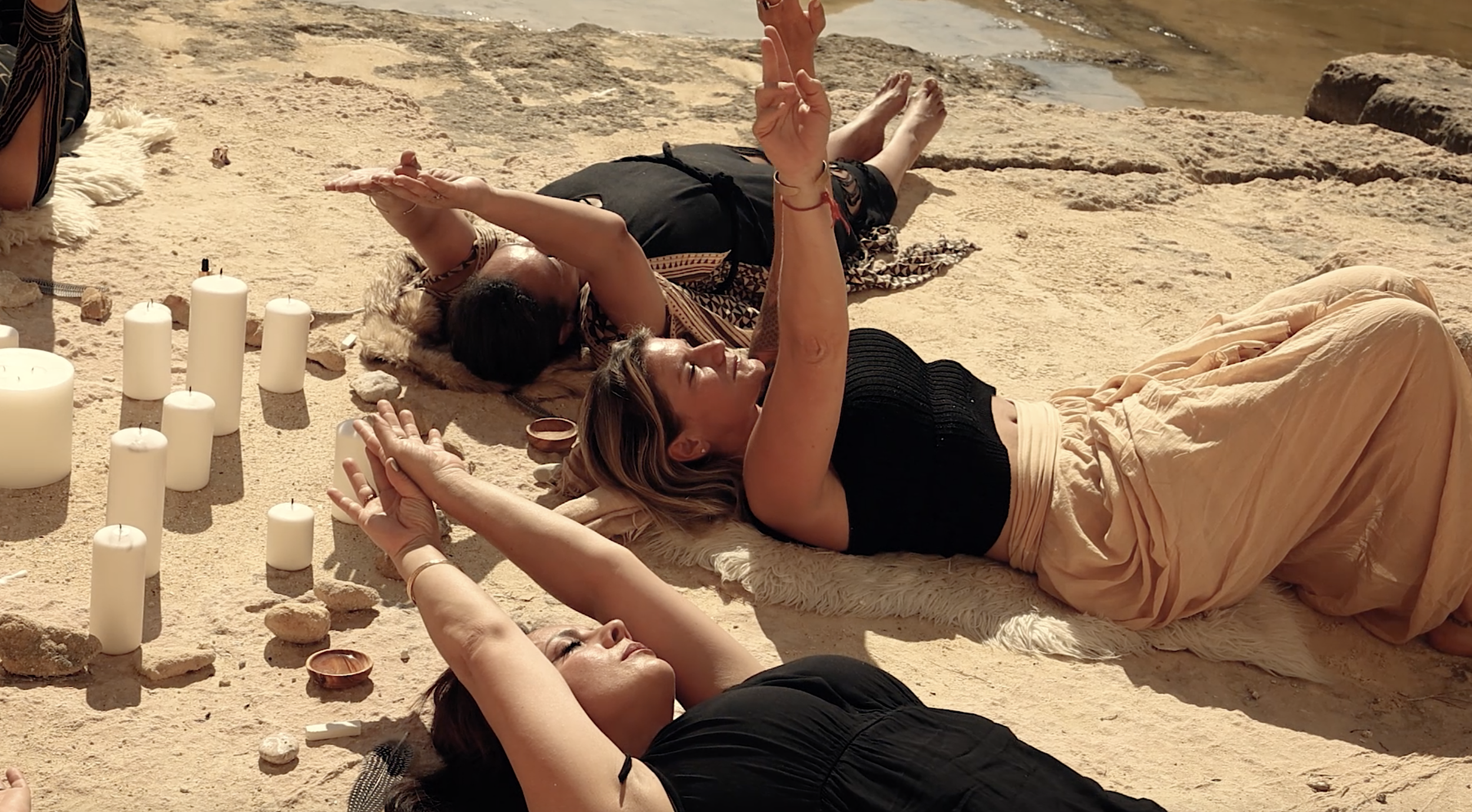 Three women lying on the sand near the water, participating in a relaxing activity with candles and stones around them.