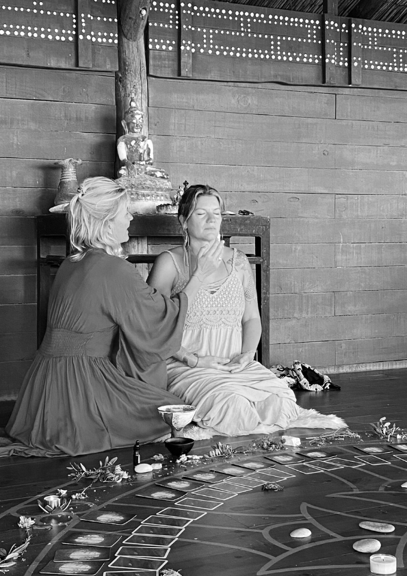 Two women participating in a spiritual or ritual ceremony indoors, with one woman applying a mark on the other's forehead. The setting has wood paneling, statues, and ritual objects, including decks of cards and decorative items on the floor.