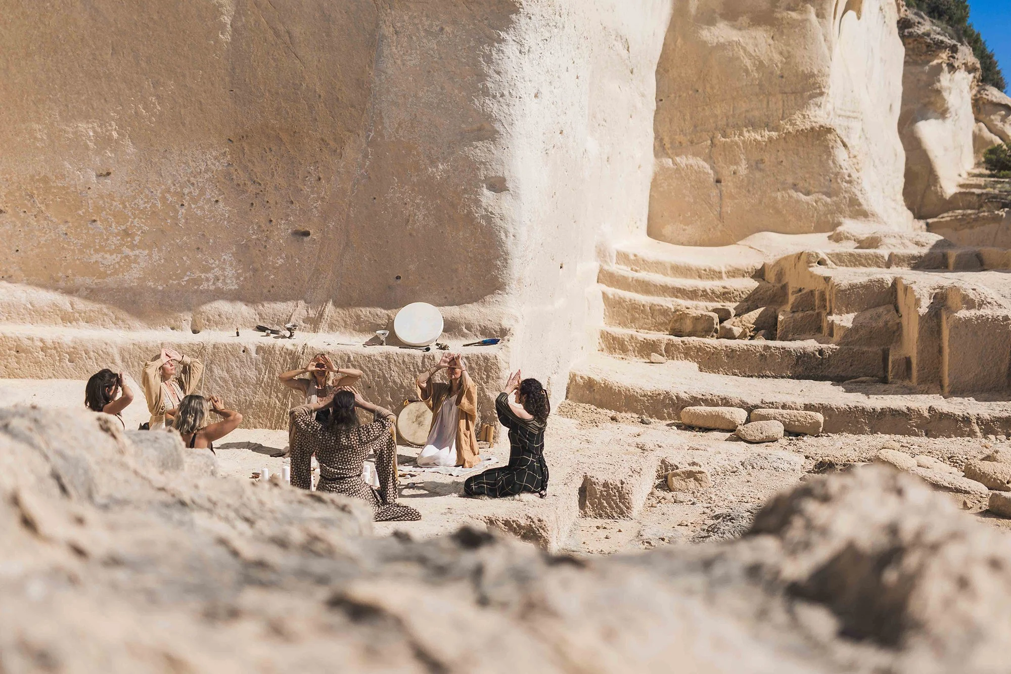 A group of six women sitting and kneeling on the ground inside an ancient stone structure, with large steps leading up the wall and pigeons perched nearby, during daytime.