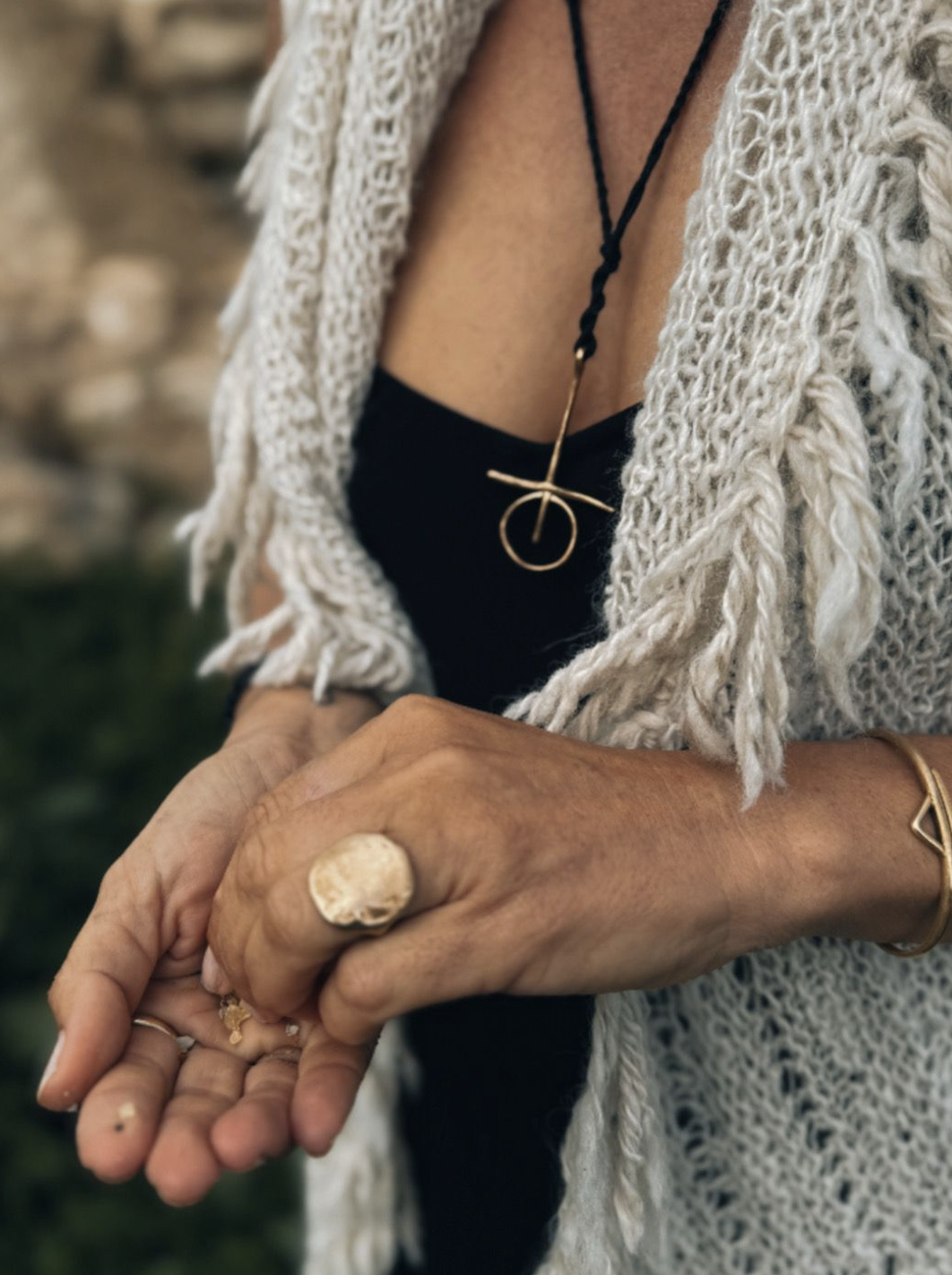 Close-up of a person's hands holding out their palm, wearing multiple rings, with a large ring on the middle finger. The person is dressed in a black top and a chunky knit white vest with fringes. They are also wearing layered jewelry, including a distinctive gold necklace with a circular pendant and a bracelet with a triangle charm. The background appears to be outdoors with greenery and a stone wall.