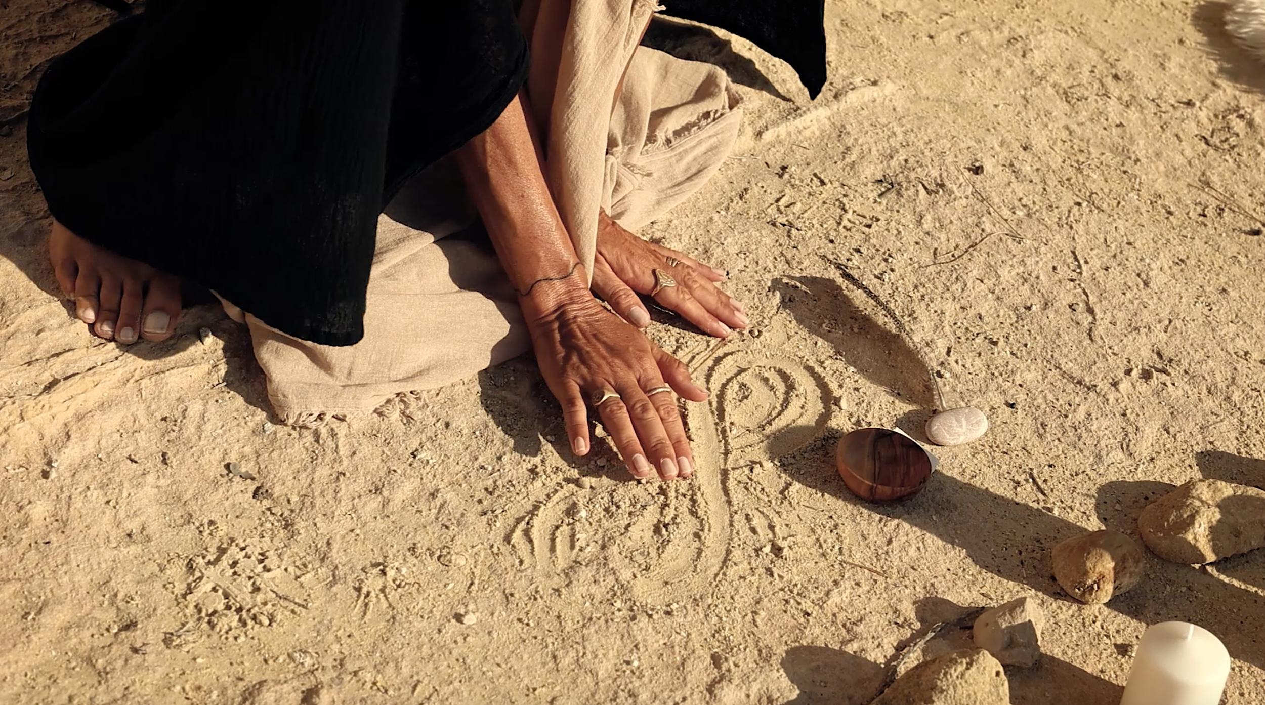 Person drawing a spiral and abstract design in the sand on a beach, surrounded by stones and a candle.