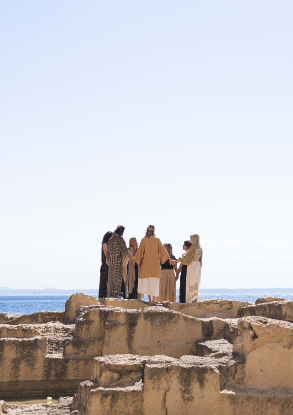 Group of women holding hands in a circle on a rocky beach with ocean in the background.