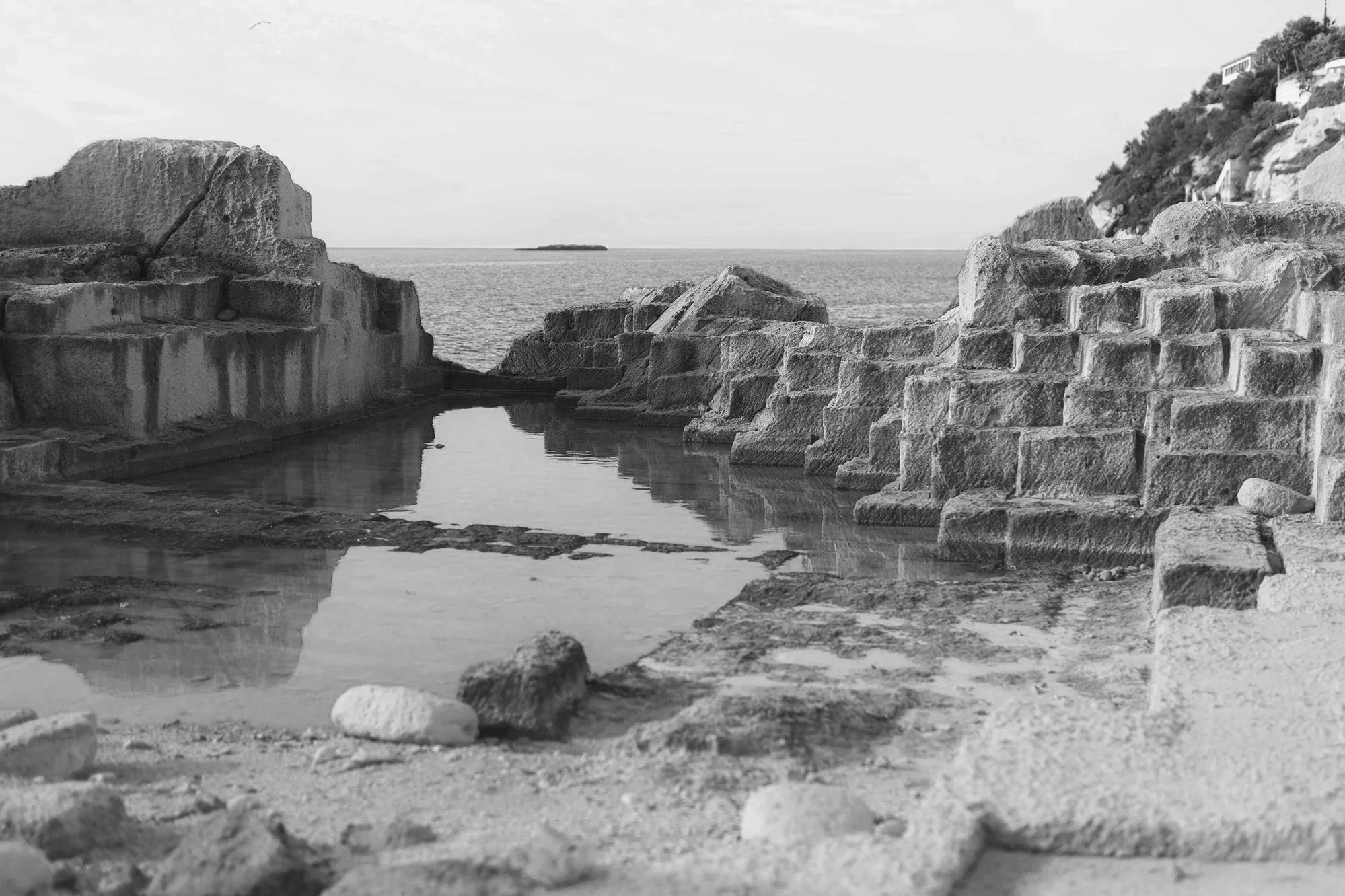 Black and white photo of a rocky beach with a small pool of water, large rocks, and a coastline with buildings on a hill in the distance.