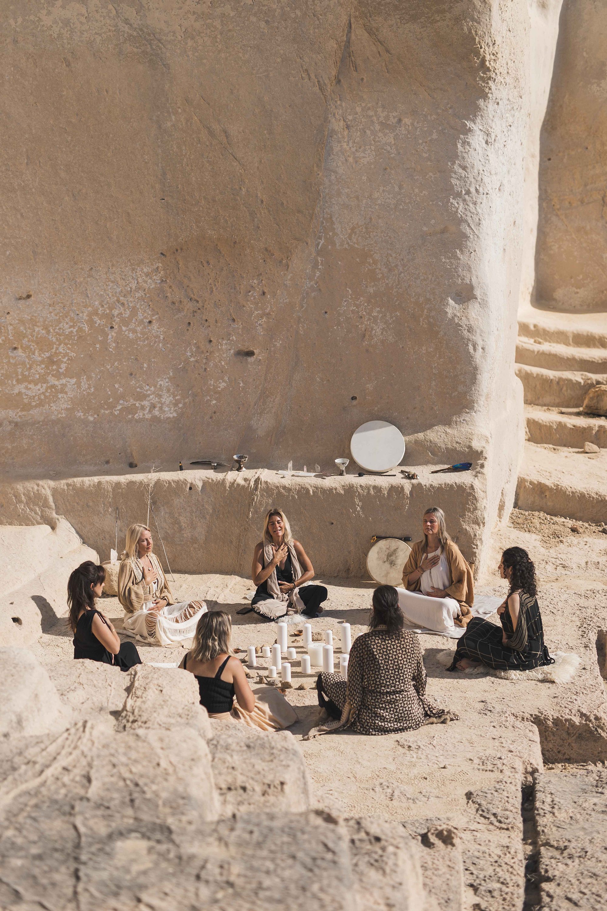 Group of women sitting in a circle inside a cave, with candles and mirrors on the ground and large round stones around.