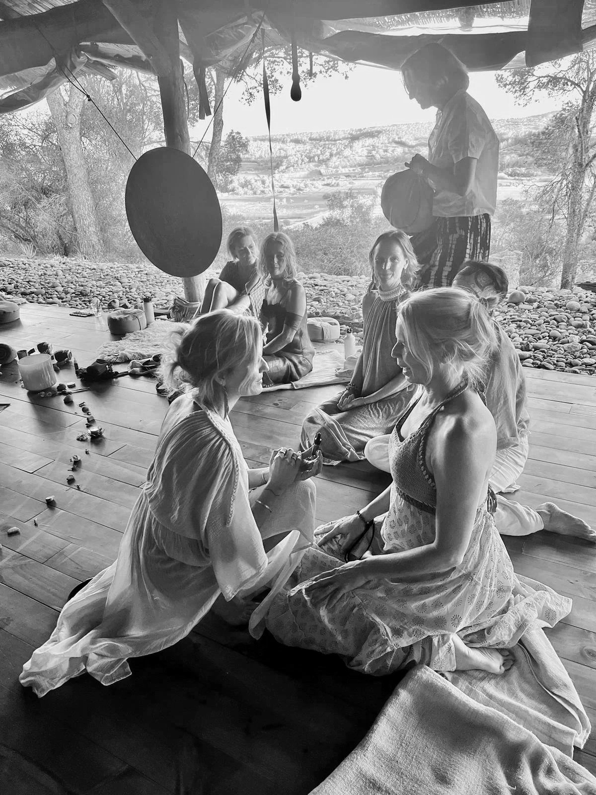 Group of women participating in a spiritual or meditation session outdoors on a wooden deck with a scenic landscape in the background, some sitting and some kneeling, with items such as candles and stones in front of them.