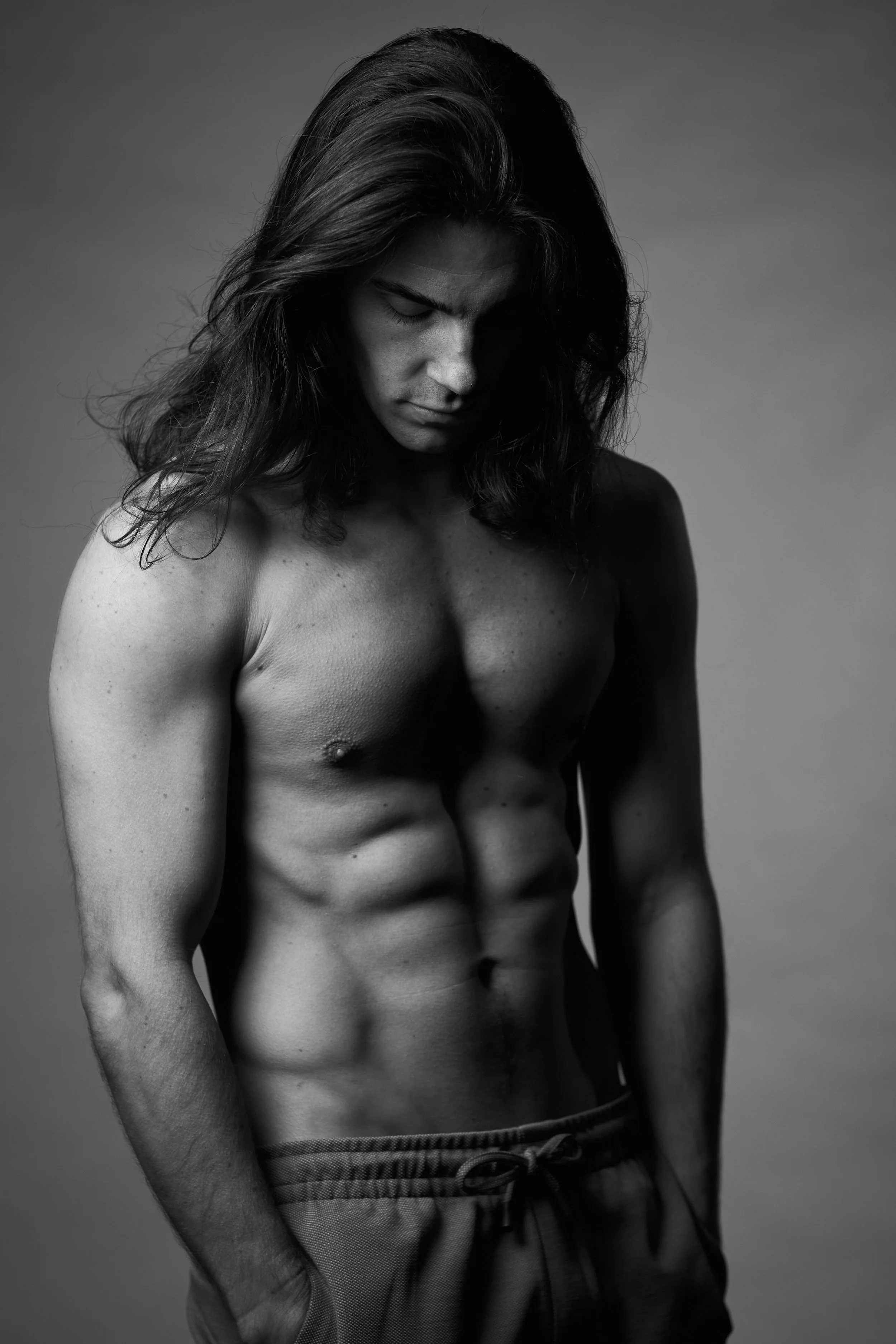 Black and white photo of a shirtless man with long hair, looking down, in a studio setting.