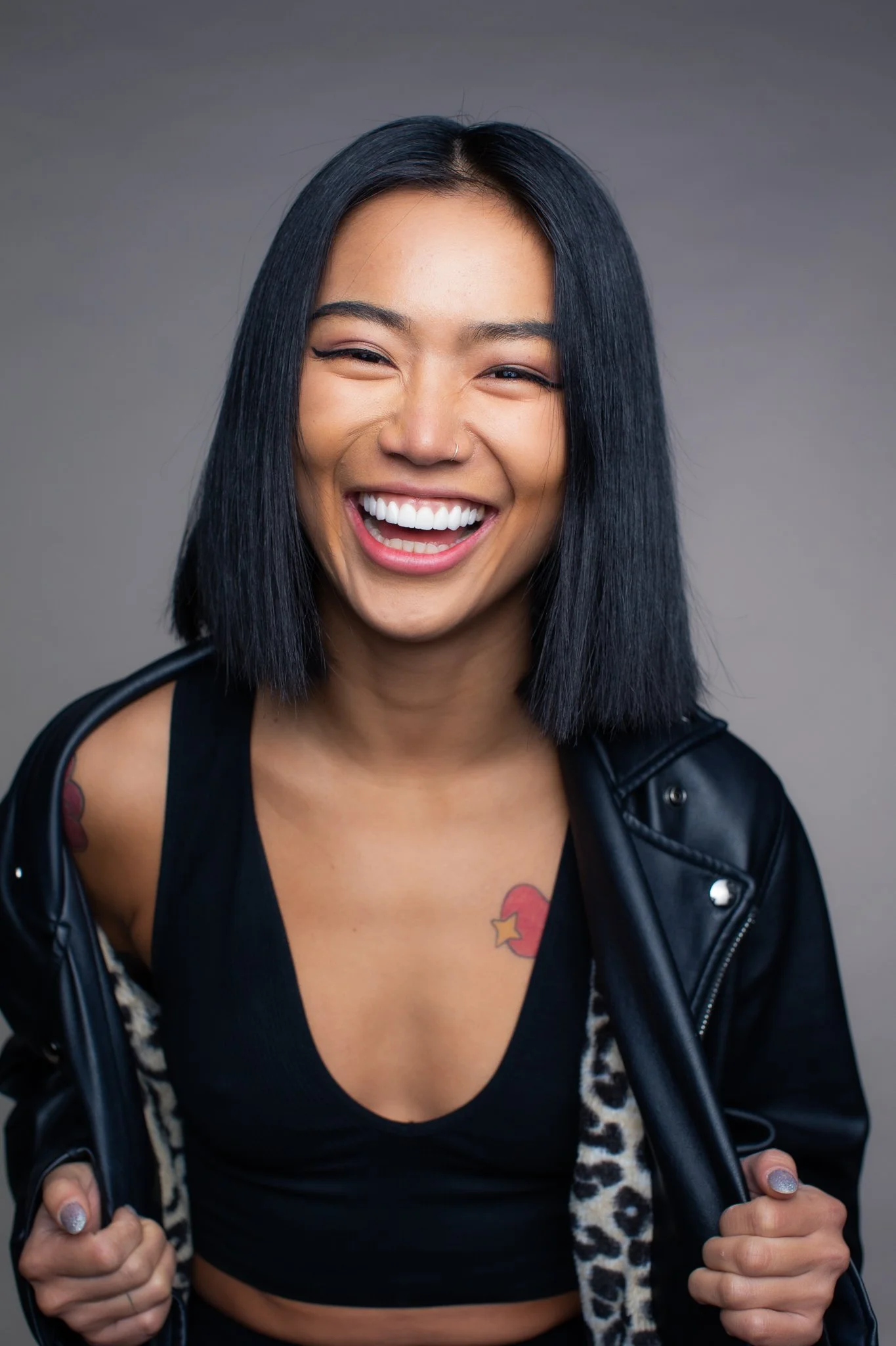 A young woman with black hair smiling, wearing a black tank top, a black leather jacket, and showing tattoos on her shoulder and chest.