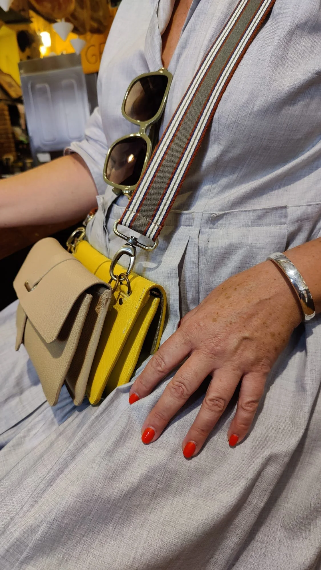 Close-up of a person dressed in a light linen dress. The person has a two Françoise City leather handbags, one yellow and another beige. The bags and a pair of sunglasses are hanging from a striped canvas strap.