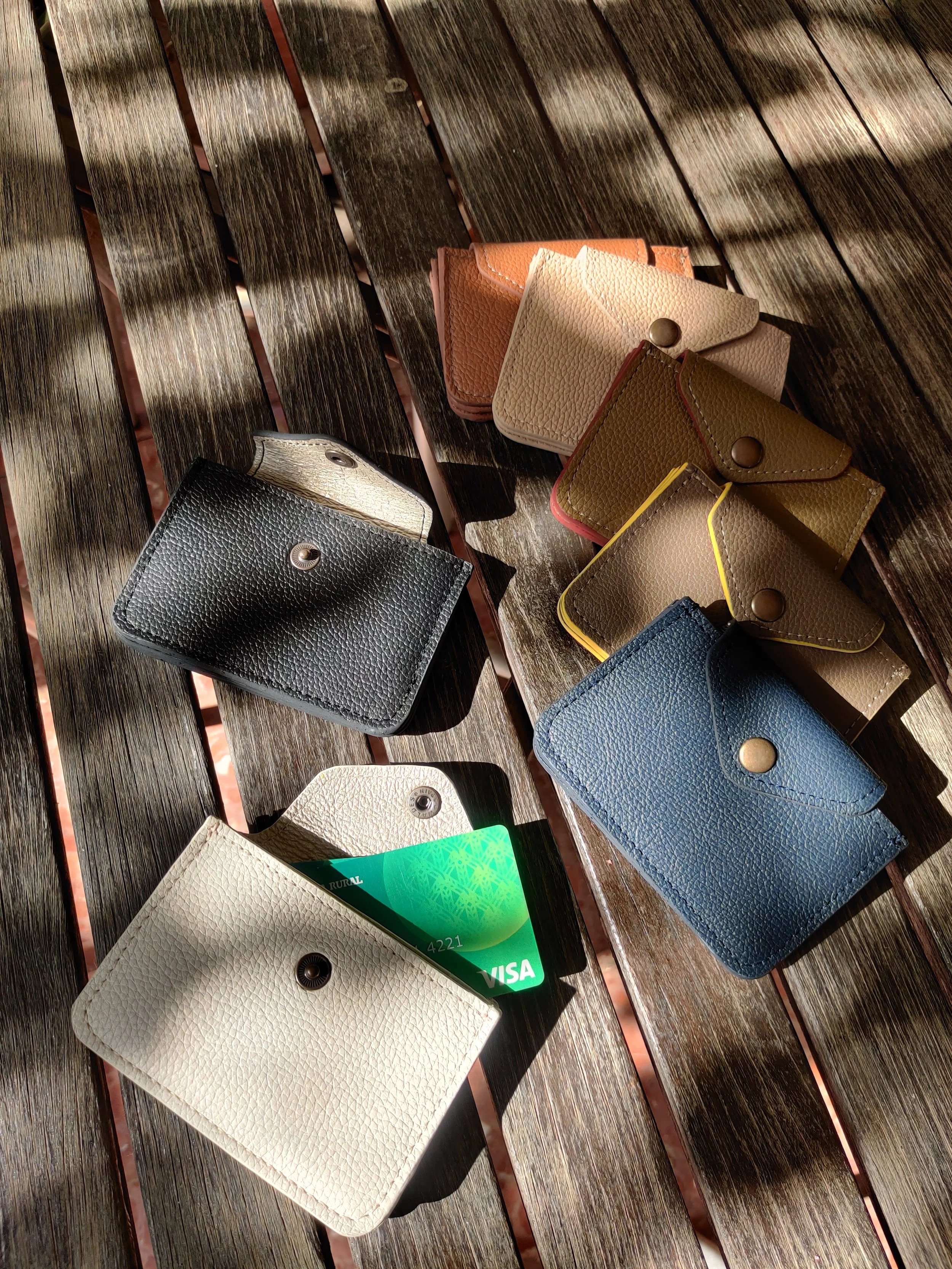 Multiple small leather cardholders of various colours, some with cards inside, are laid out on a wooden surface with sunlight and shadows.