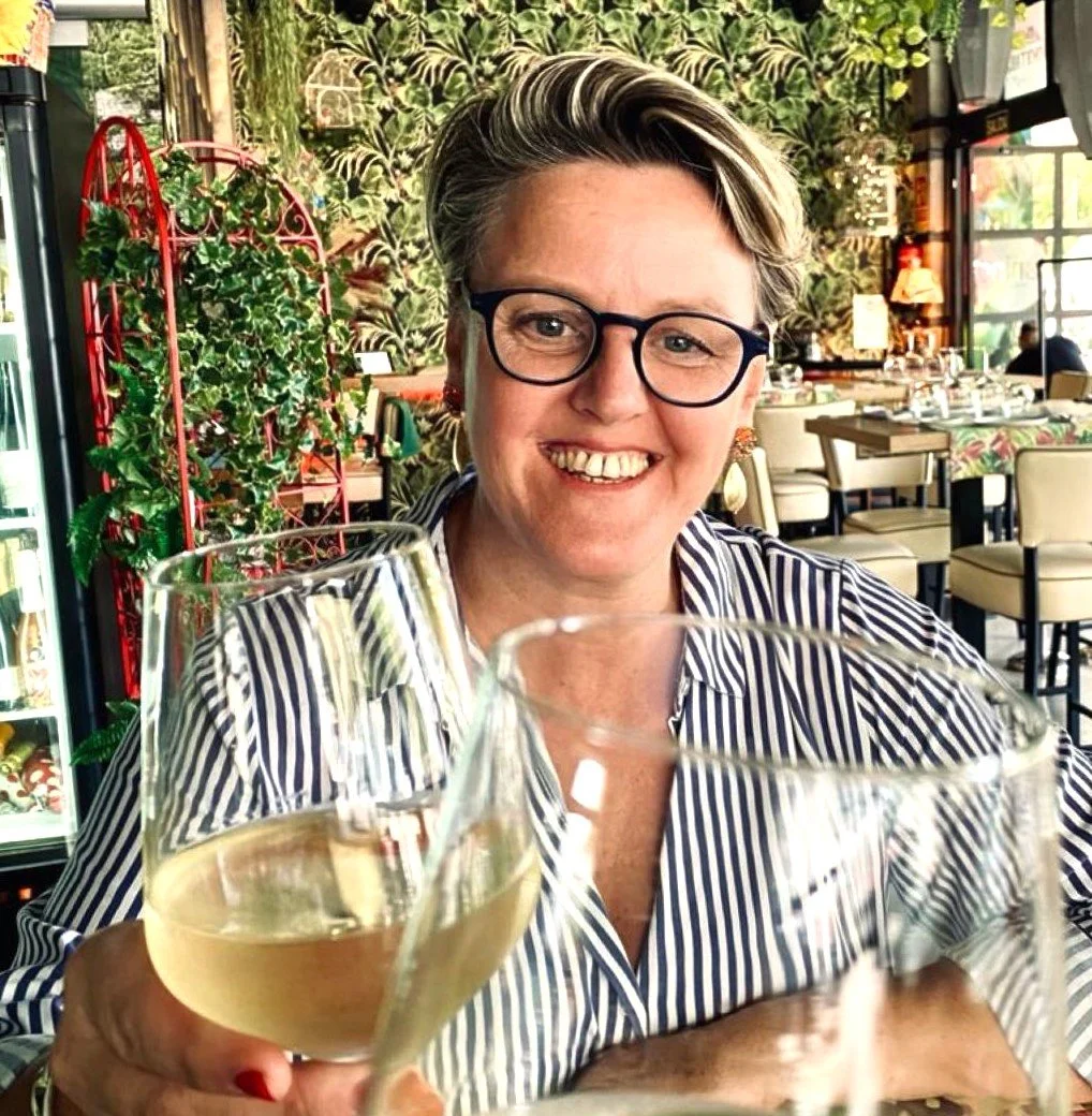Francoise, the owner of Boutique Unique, with short blonde hair, glasses, and a striped shirt smiling while holding a glass of white wine inside a restaurant decorated with plants.