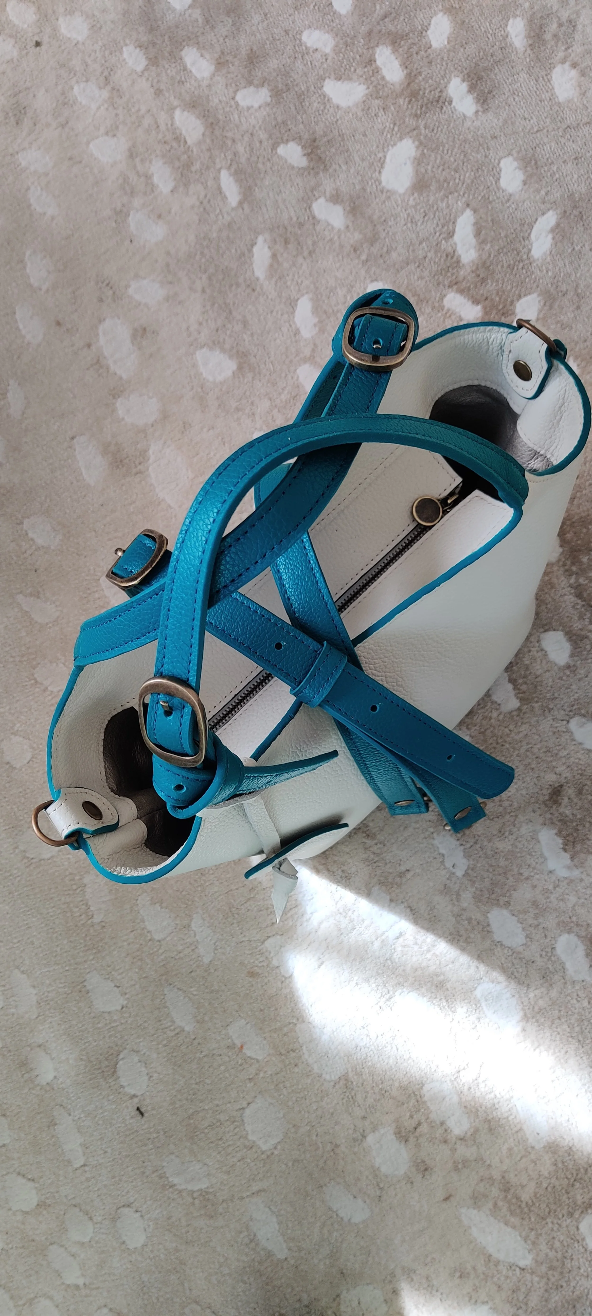 A white handbag with blue straps and metal buckles, placed on a beige patterned surface.