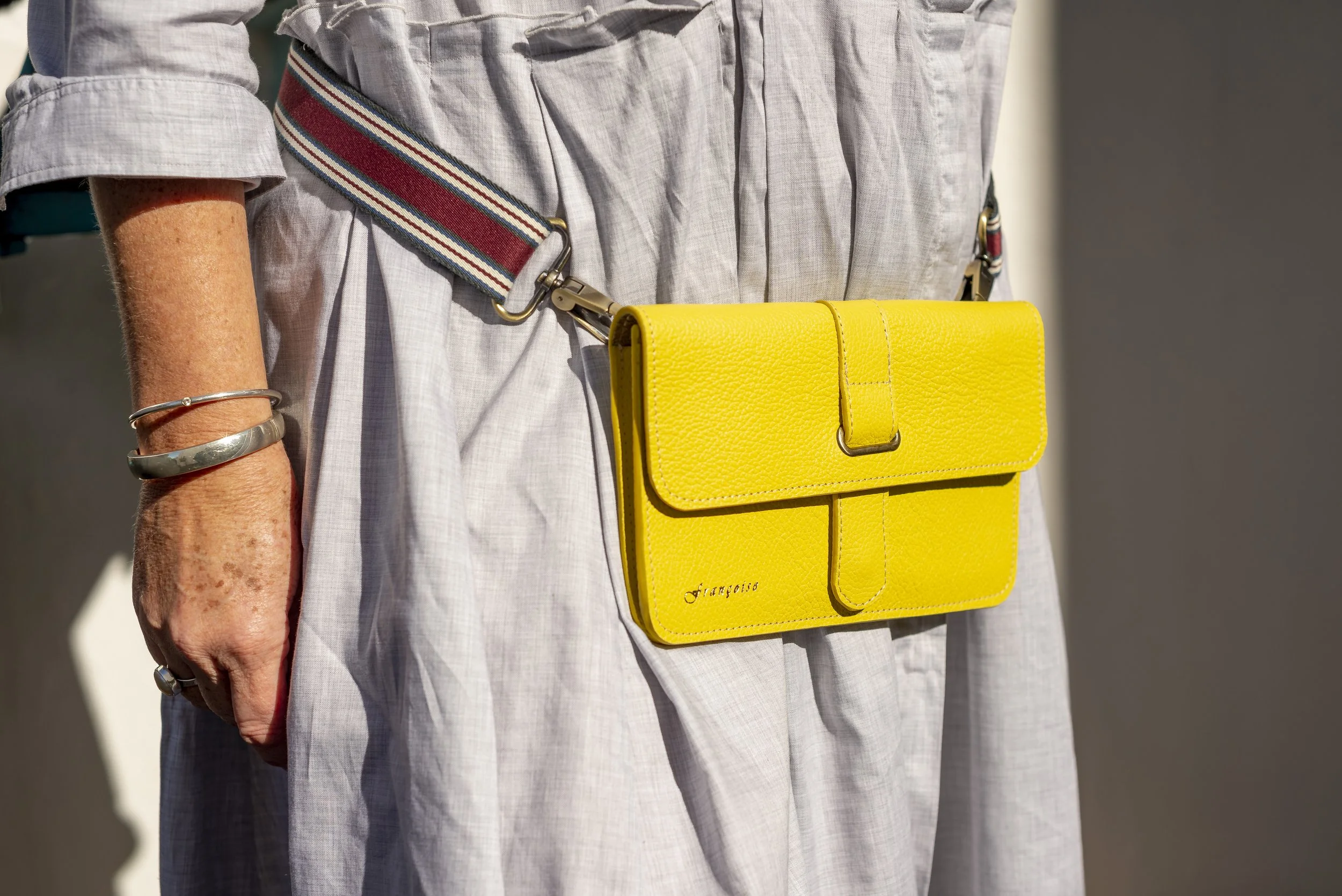 Close-up of a person wearing a light-colored linen dress with rolled-up sleeves, a bracelet on their wrist, and a colorful striped strap attached to a yellow Françoise City leather handbag.