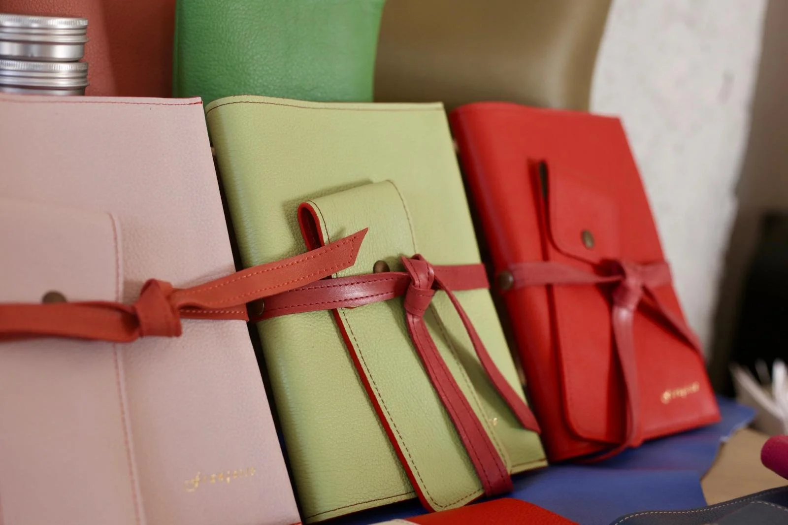 Multiple colorful notebooks with leather straps, arranged on a surface, with a blurred background including a white wall and other objects.