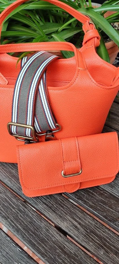 An orange handbag showing a matching wallet in front of the bag. Both have been placed on a wooden surface with green plants in the background. There is also a striped strap hanging from the handbag.